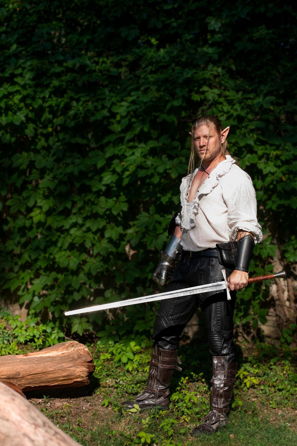 Medieval warrior in leather armor poses with sword near fallen log in forest.