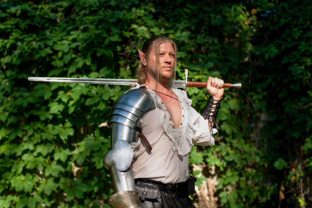 Adventurer in medieval garb wields a sword and drinking vessel in a wooded area.