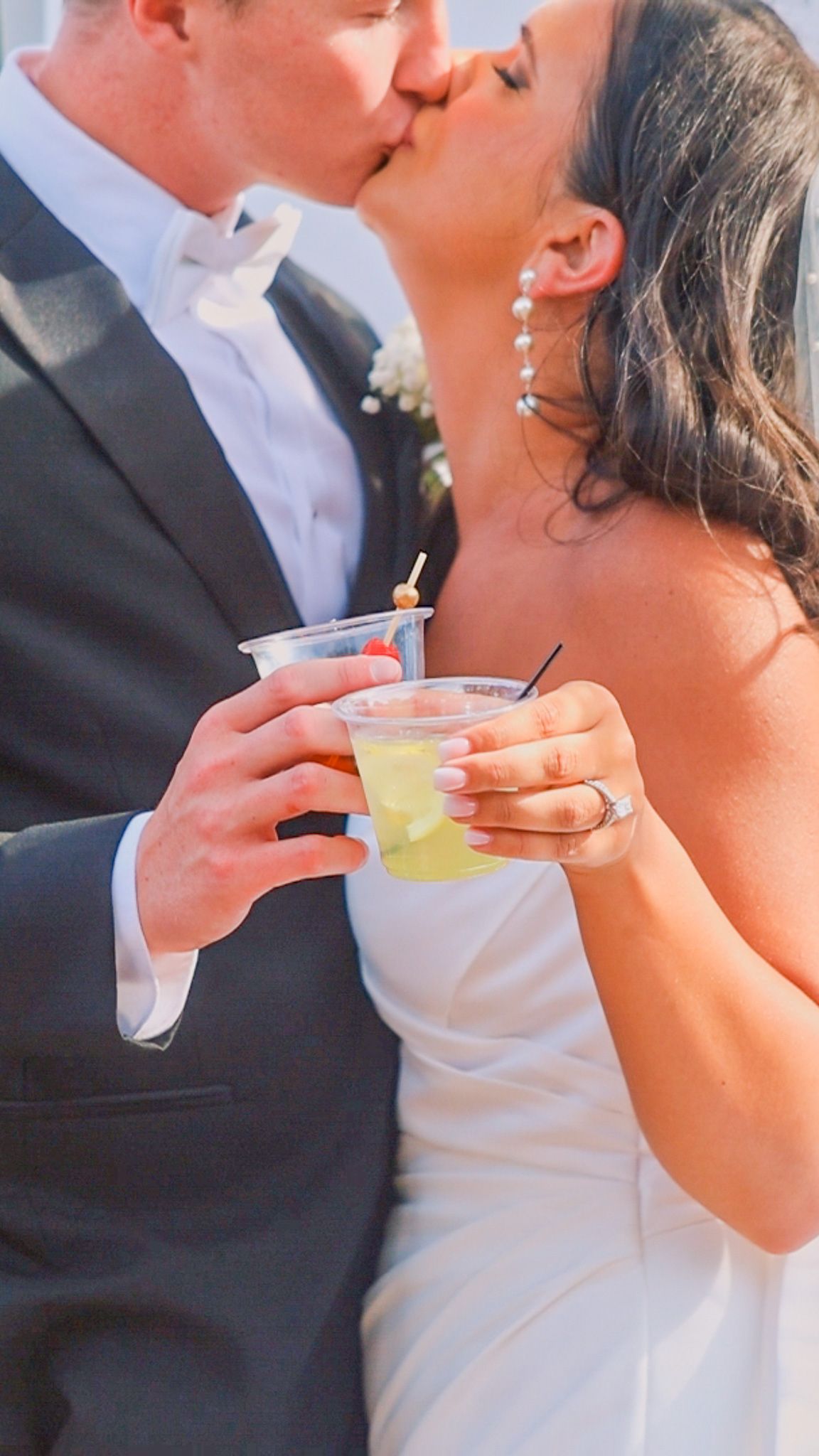 Newlyweds share a kiss while holding cocktails at their wedding reception, with bride in white and groom in black suit.