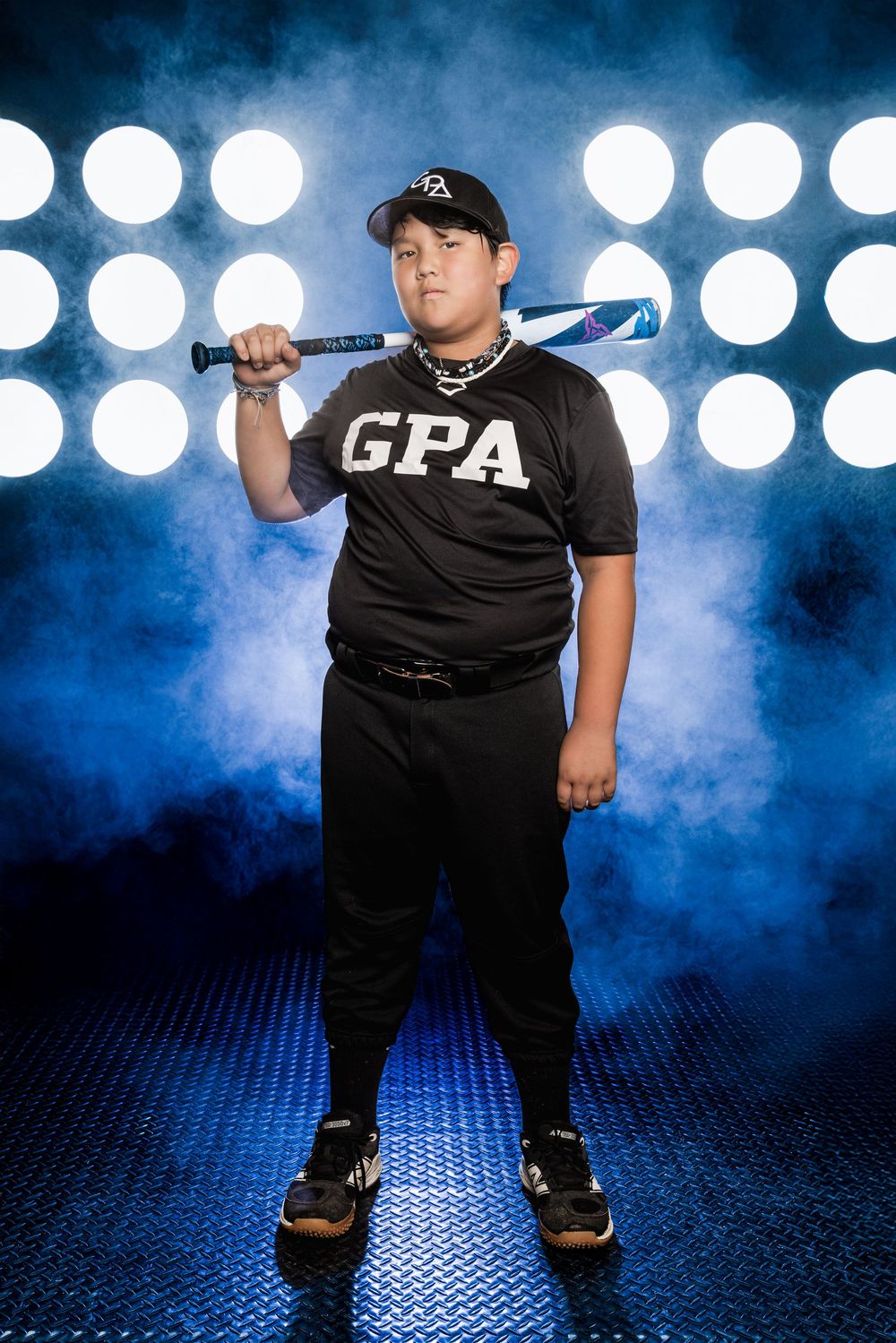 Performer in black clothing and baseball cap poses dramatically against blue spotlight backdrop on textured stage floor.