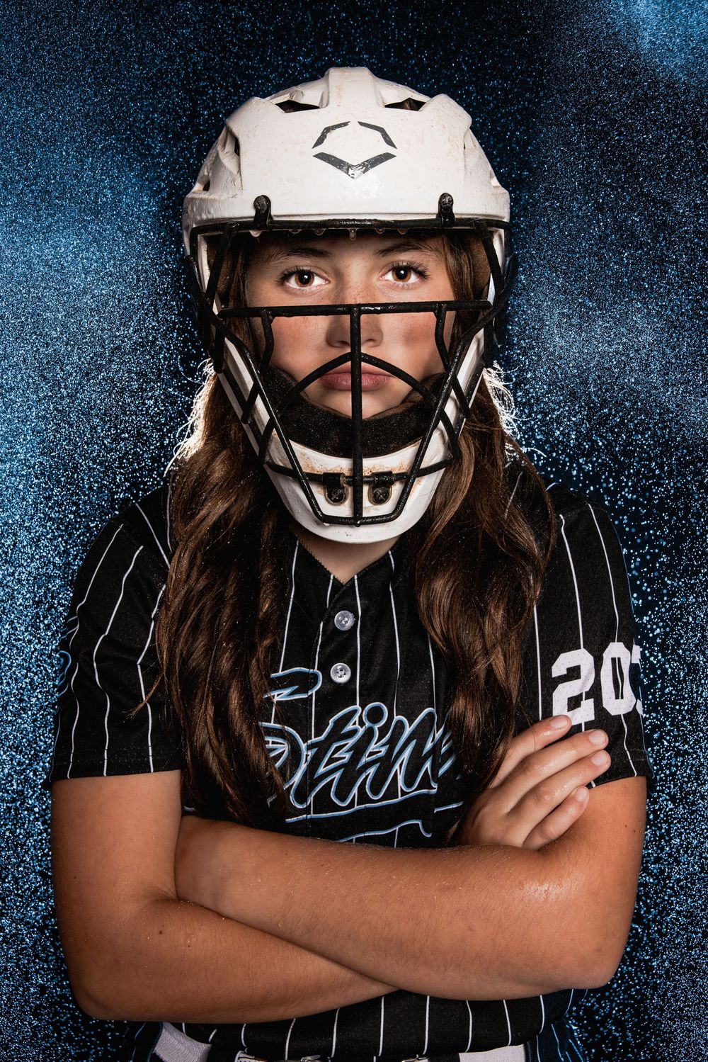Softball player in black pinstripe uniform and protective helmet poses with crossed arms in dramatic lighting.