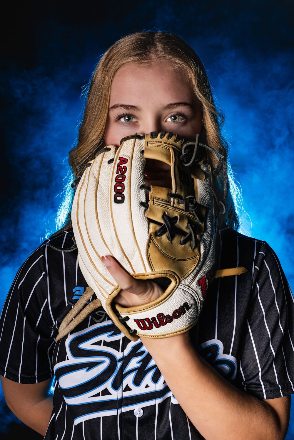 Softball player in pinstripe jersey holds white and tan glove in front of face against dramatic blue smoke background.