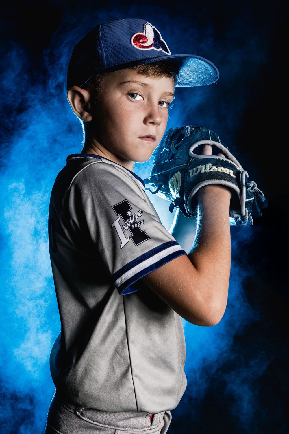 A youth baseball player poses in uniform with a Wilson glove against a dramatic blue smoke background.