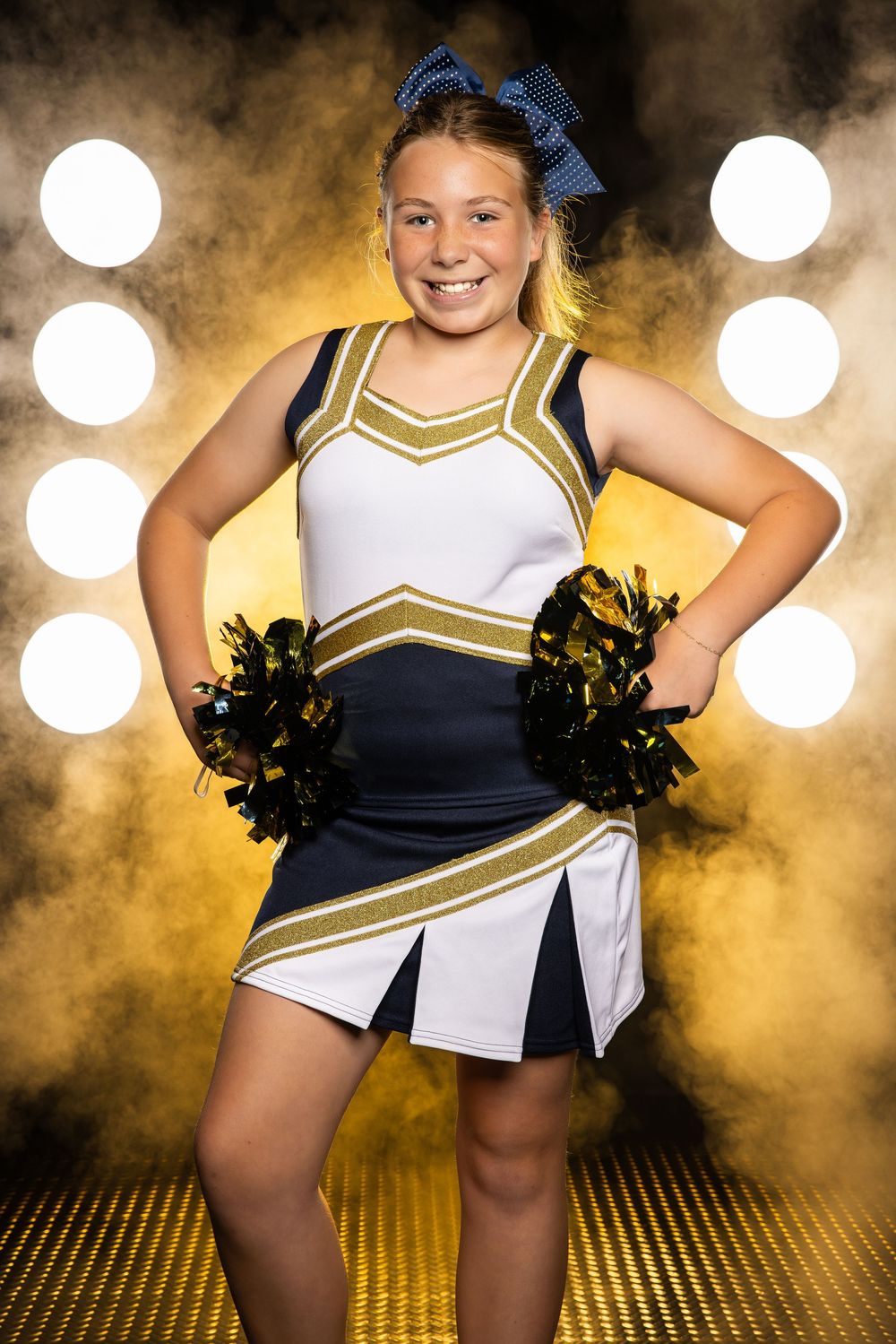 Cheerleader in navy and gold uniform strikes dynamic poses with pom poms against dramatic spotlit background.