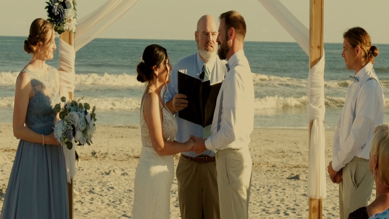A seaside wedding ceremony takes place under a wooden arch on a sunny beach.