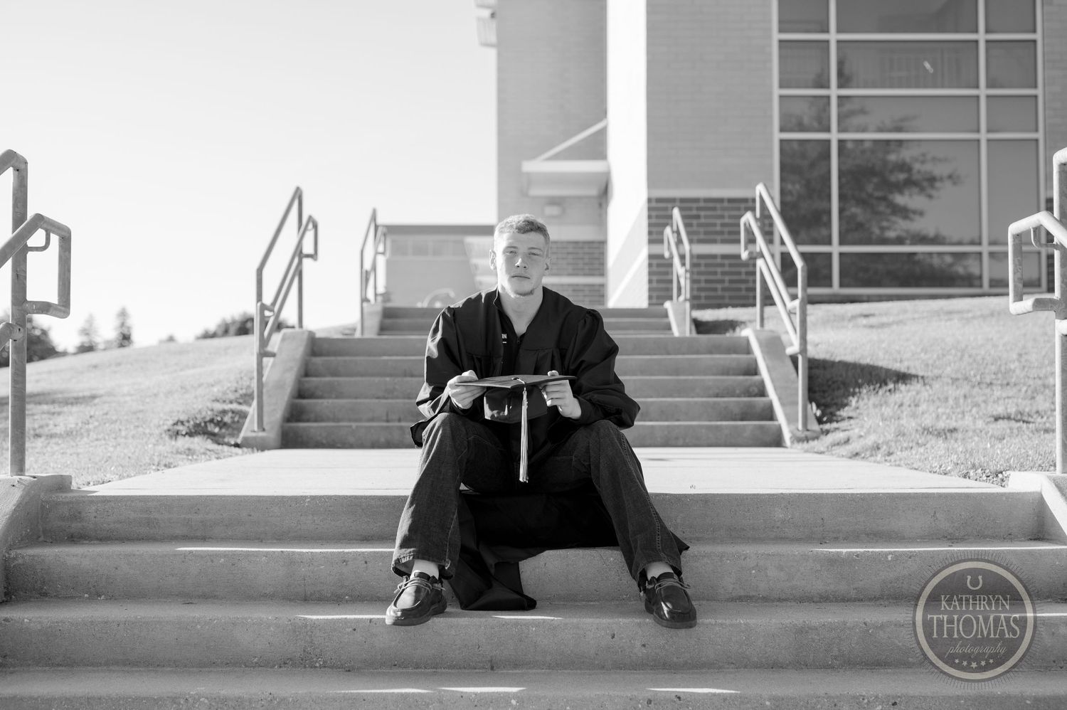 Black and white photo of graduate seated on outdoor campus steps in full regalia.