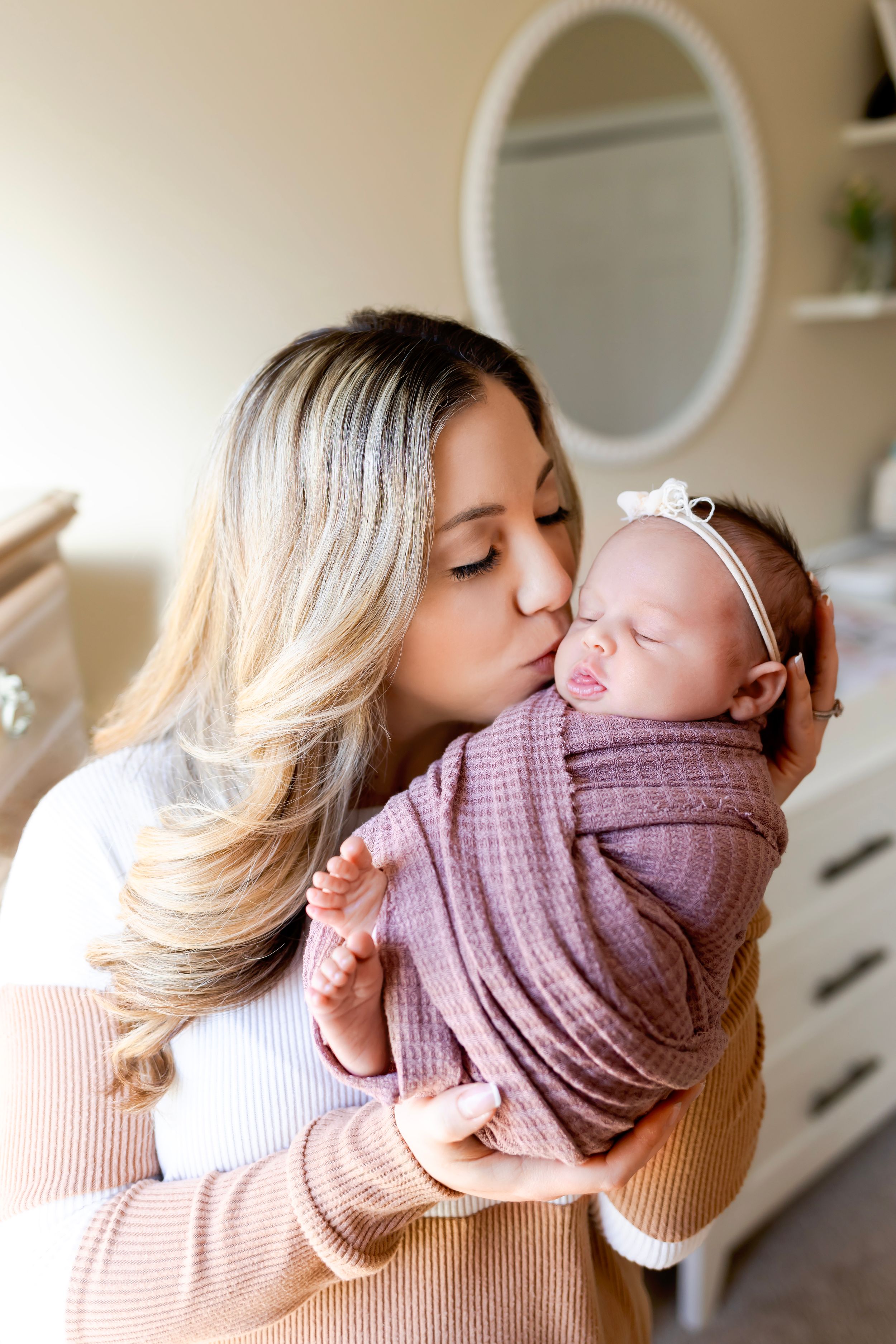 A tender moment as a woman in a beige sweater kisses a swaddled newborn baby. NJ Newborn Photographer in home session.