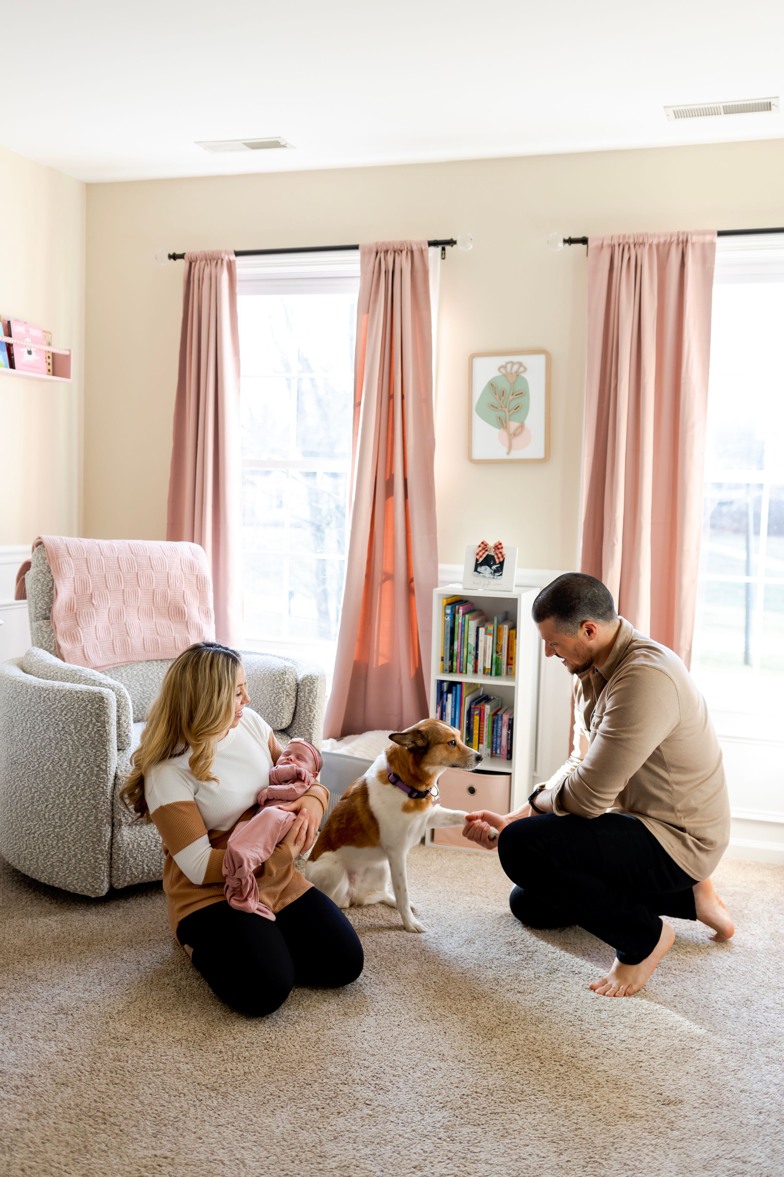 A family relaxes together in a cozy living room with pink curtains while playing with their white and brown dog. NJ Newborn Photographer
