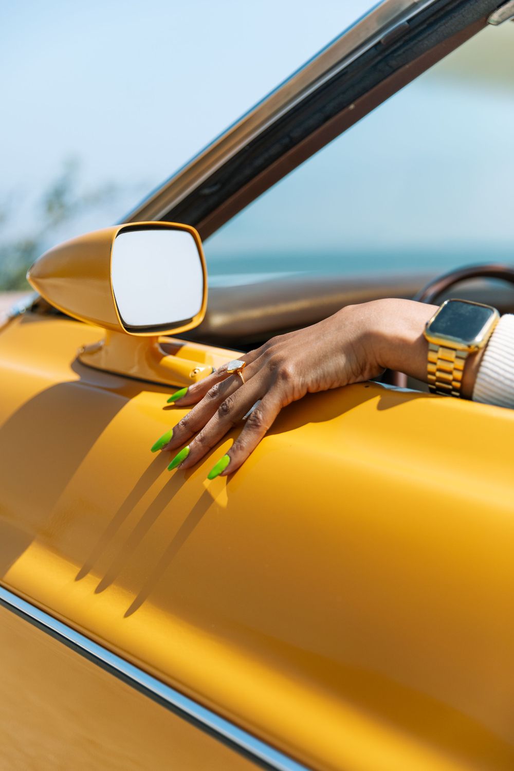 A hand wearing a gold watch rests on a shiny yellow car hood next to a side mirror in bright sunlight.