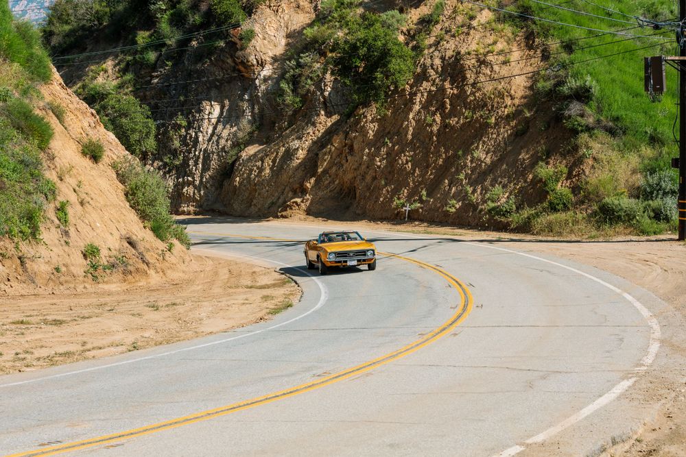 Yellow vintage muscle car navigates a winding mountain road with steep cliff walls and double yellow lines.