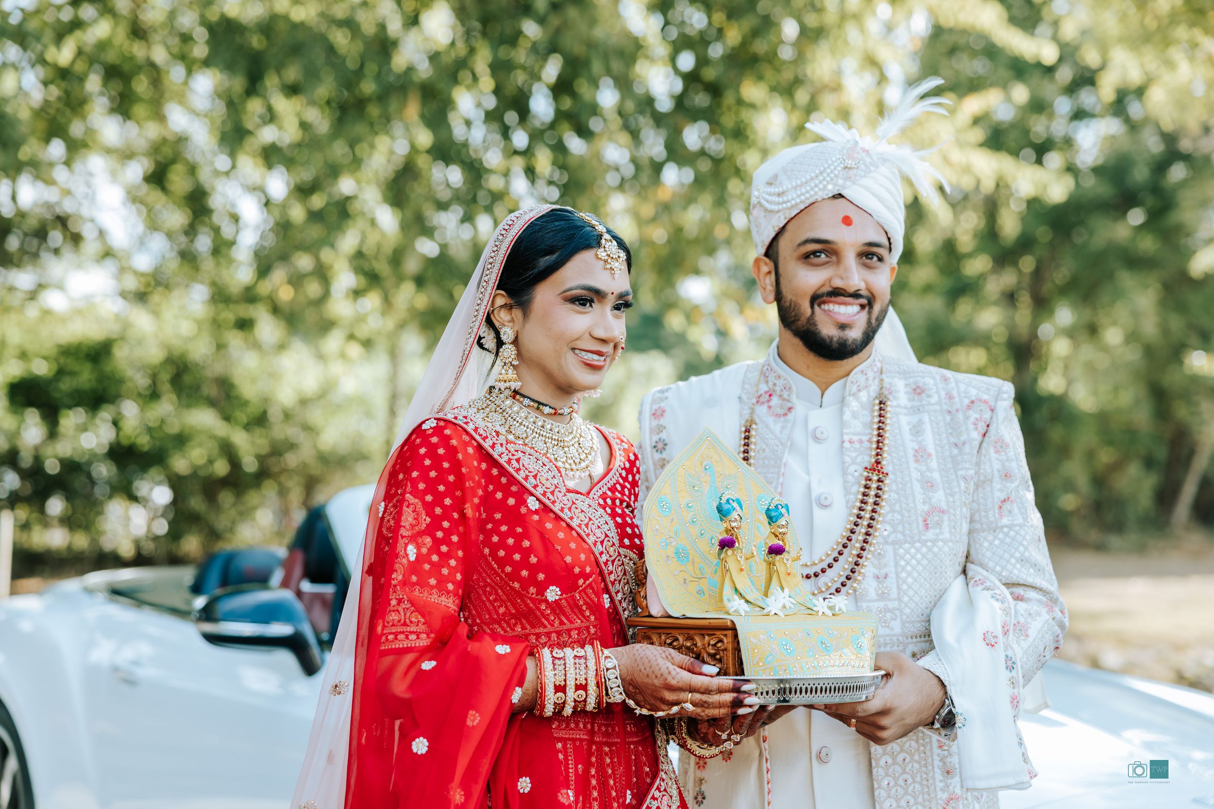 Gujarati Indian bride in red lehenga and groom in white sherwani holding a decorative tray outdoors on their wedding day.