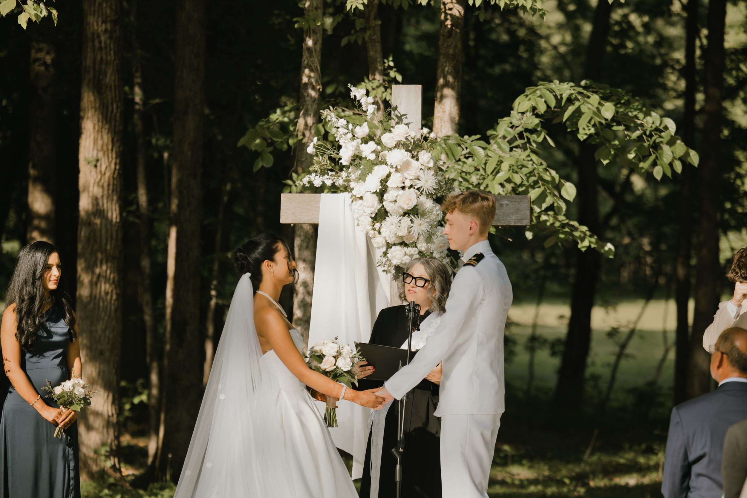 Bride and groom exchange vows outdoors in wooded setting before white cross adorned with white floral arrangements.