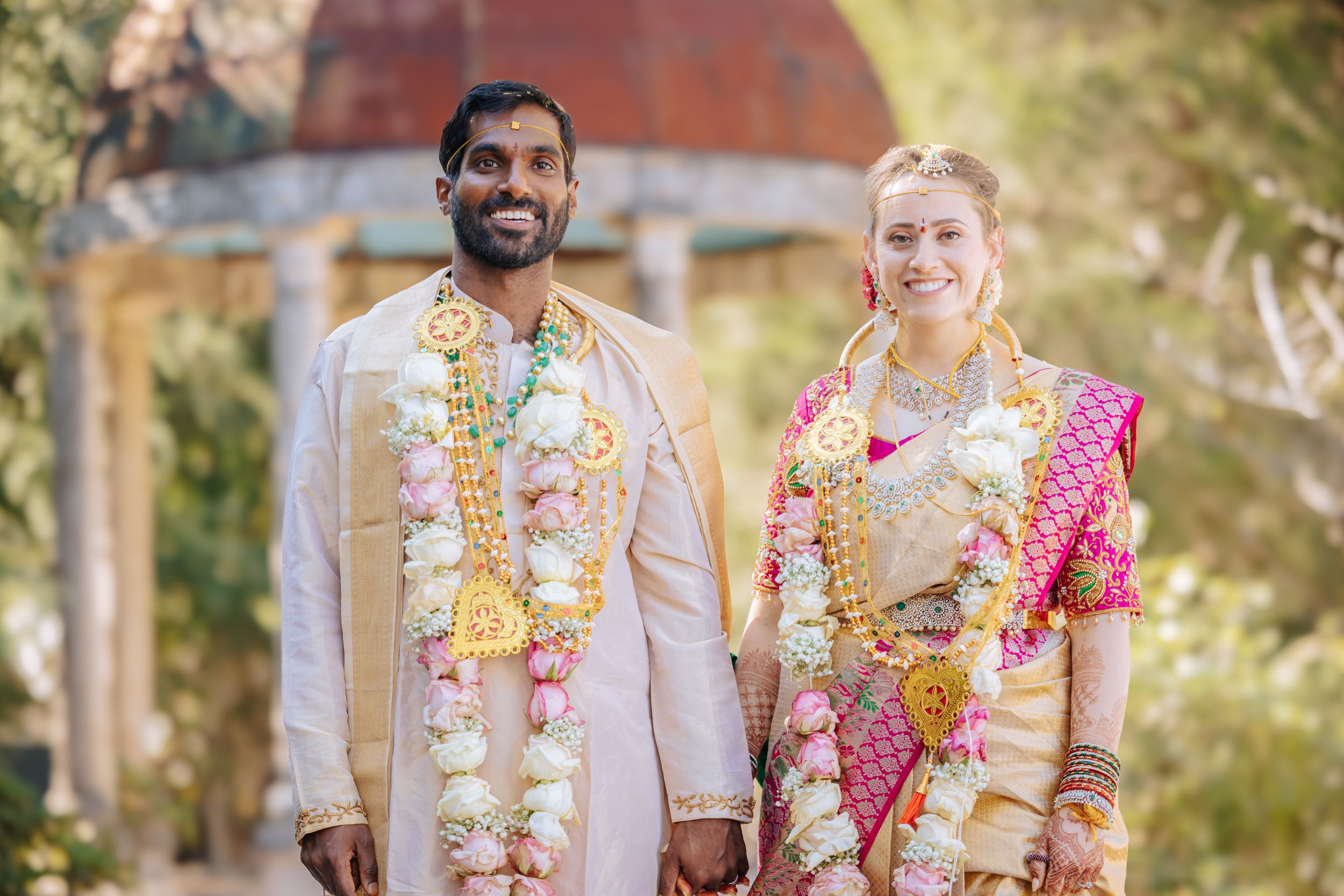 Inter-cultural Couple in traditional Indian wedding attire with floral garlands, smiling outdoors at a Hindu ceremony.