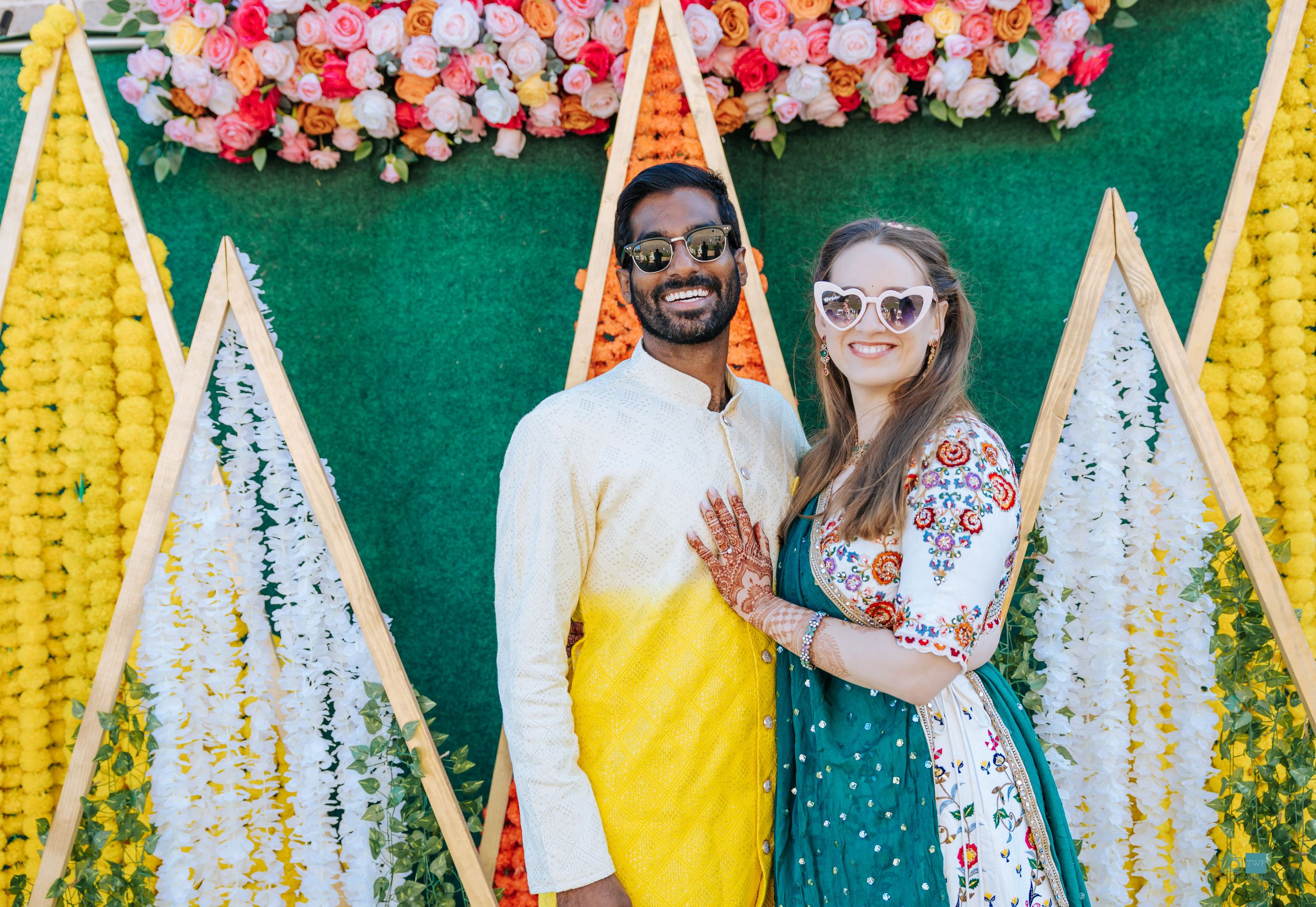 American Indian Couple posing at colorful Indian wedding backdrop with floral decorations, man in yellow kurta, woman in floral dress.