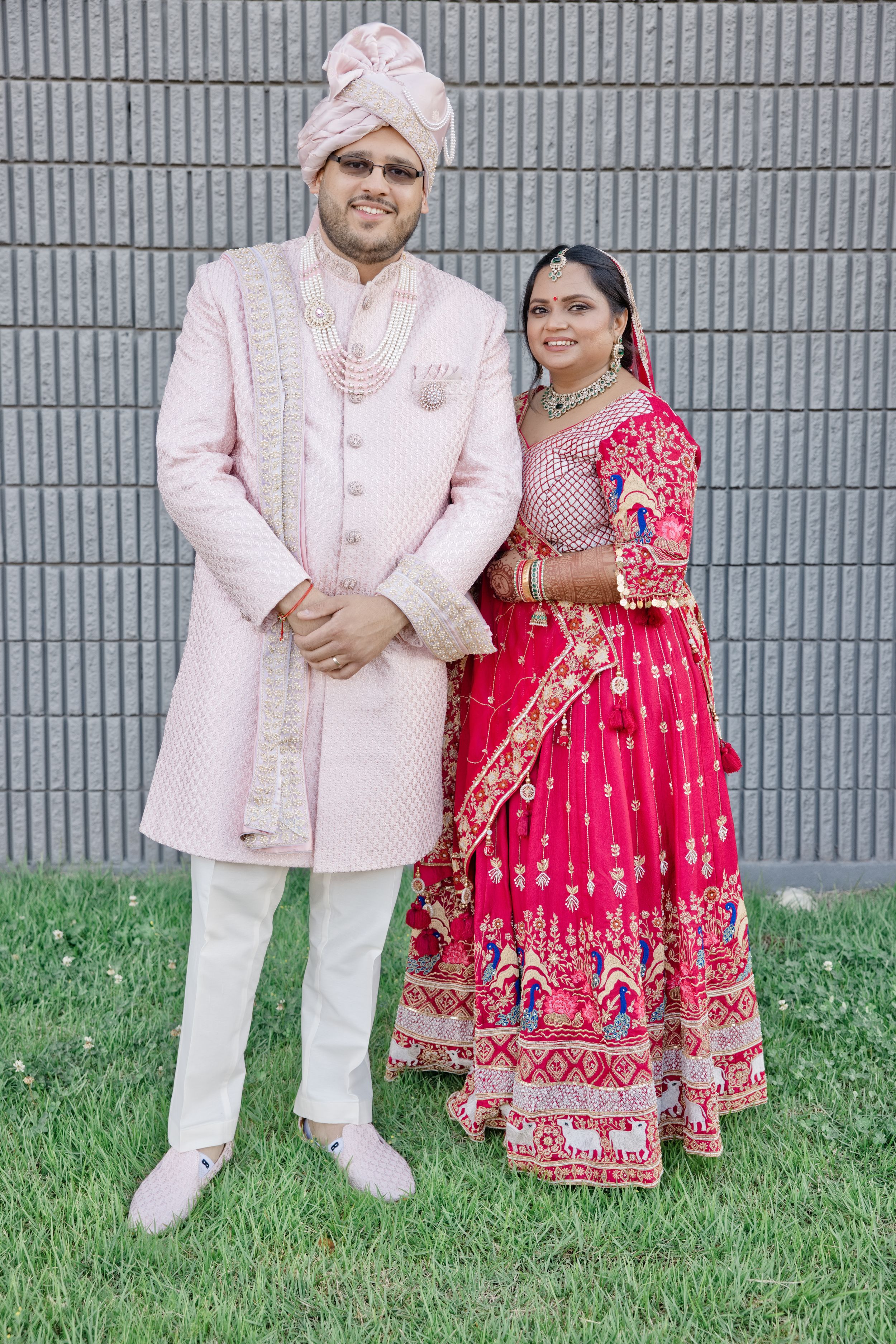 Indian couple in traditional wedding attire, groom in pink sherwani and turban, bride in red lehenga with gold embroidery.