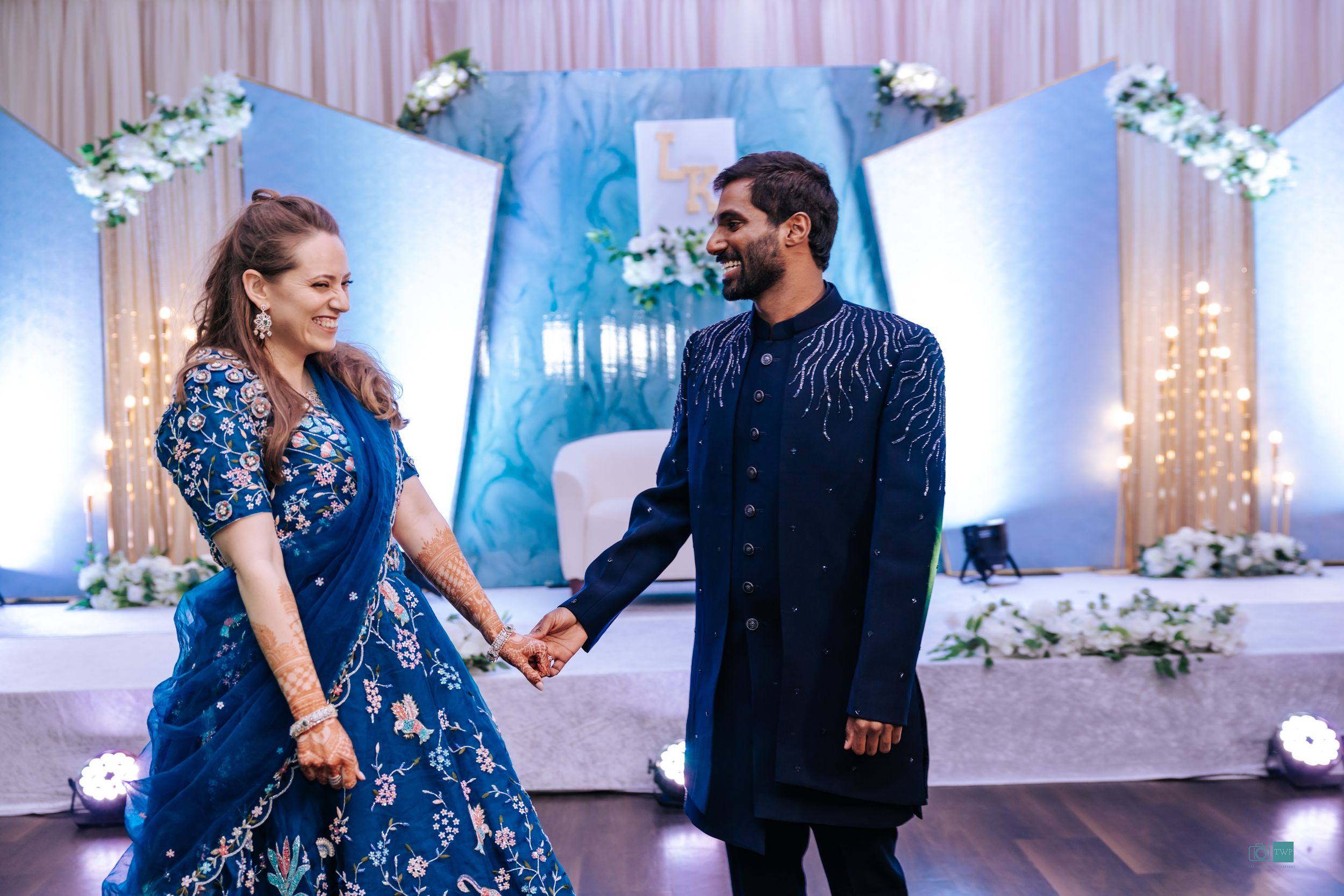 Multi-Cultural Couple in matching blue outfits holding hands at an elegant wedding reception with floral decor.