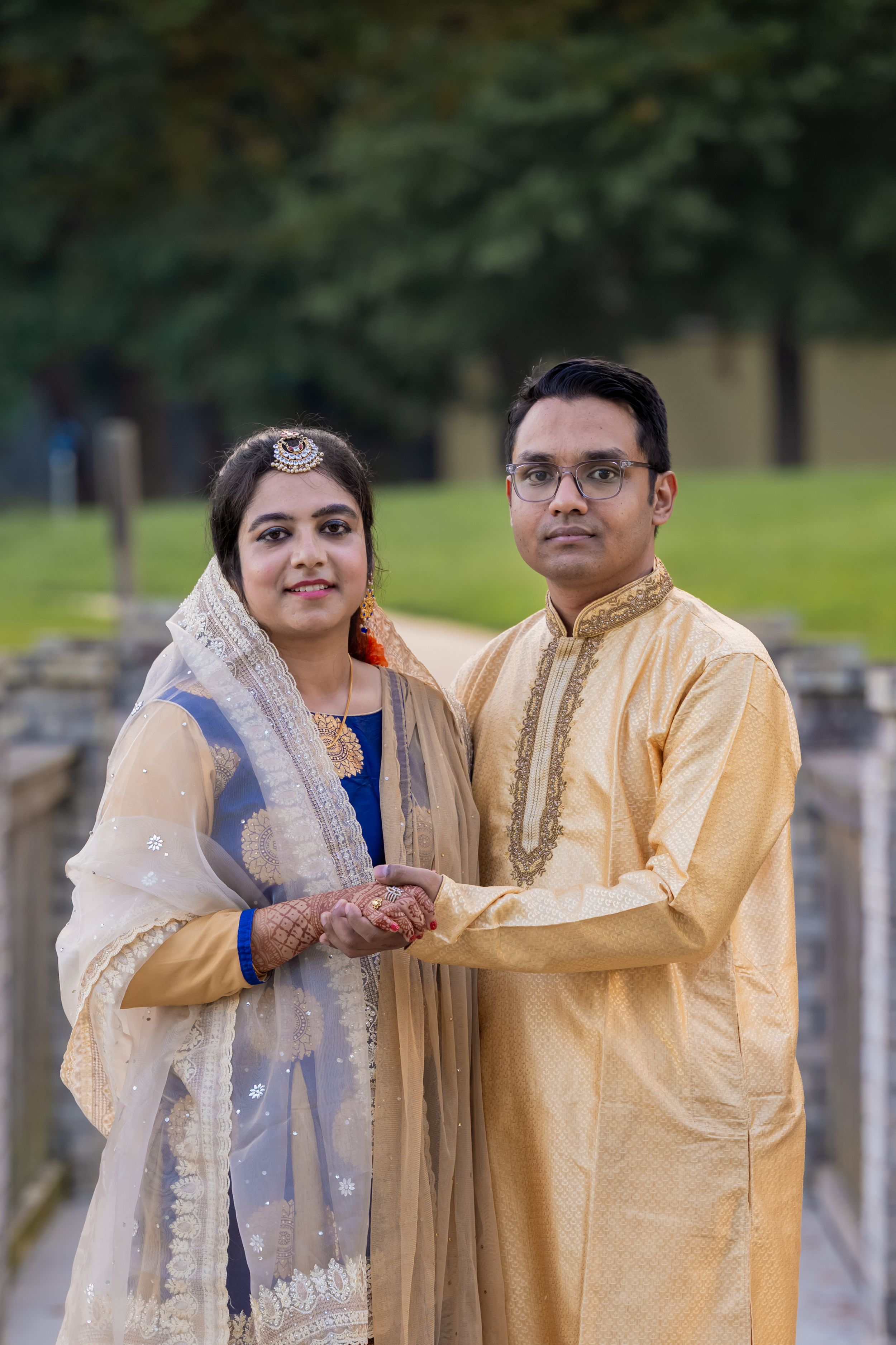 Indian couple in traditional attire holding hands outdoors, woman in blue salwar with dupatta, man in gold kurta.