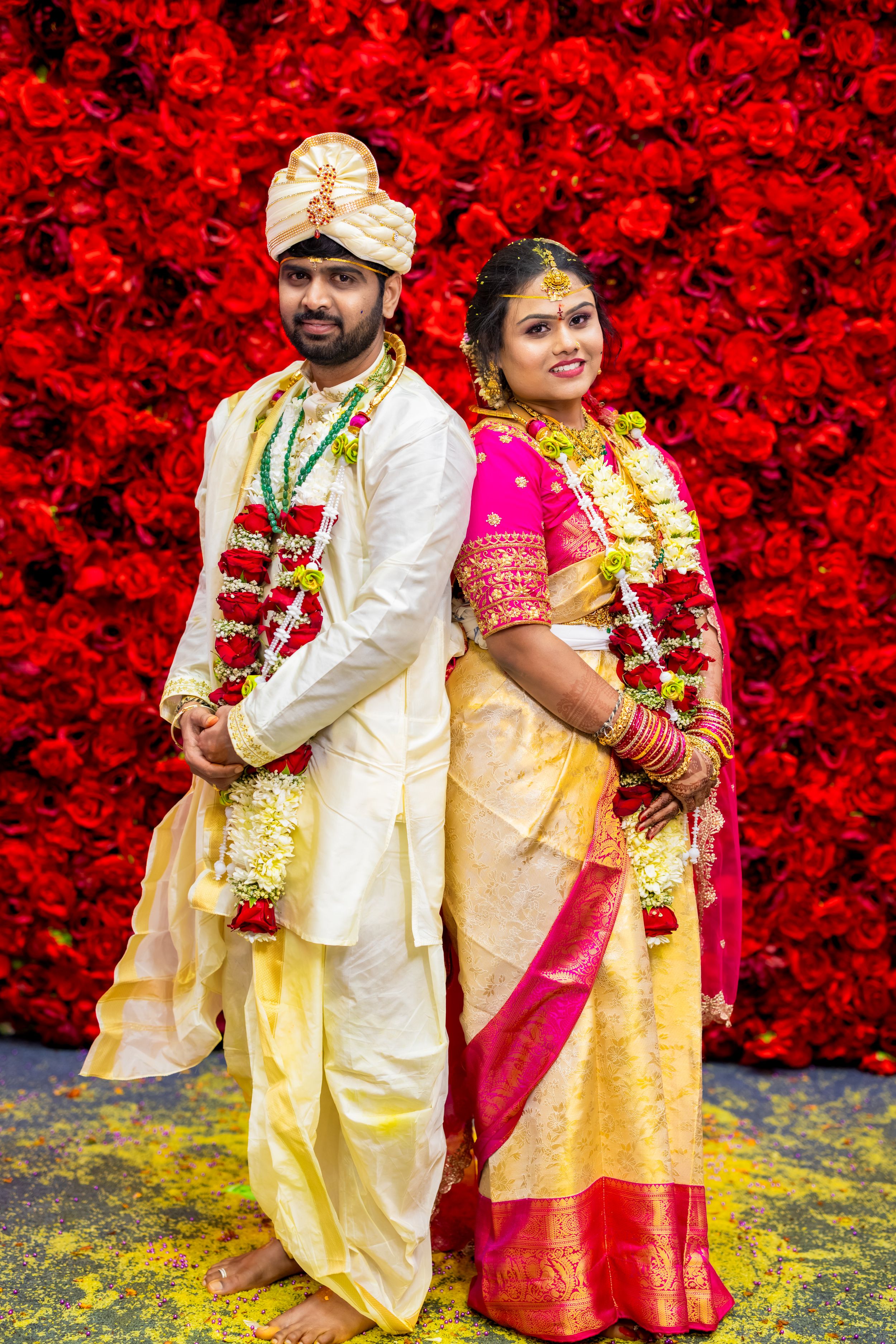 South Indian couple in traditional wedding attire pose before red floral backdrop, bride in pink saree, groom in white dhoti.