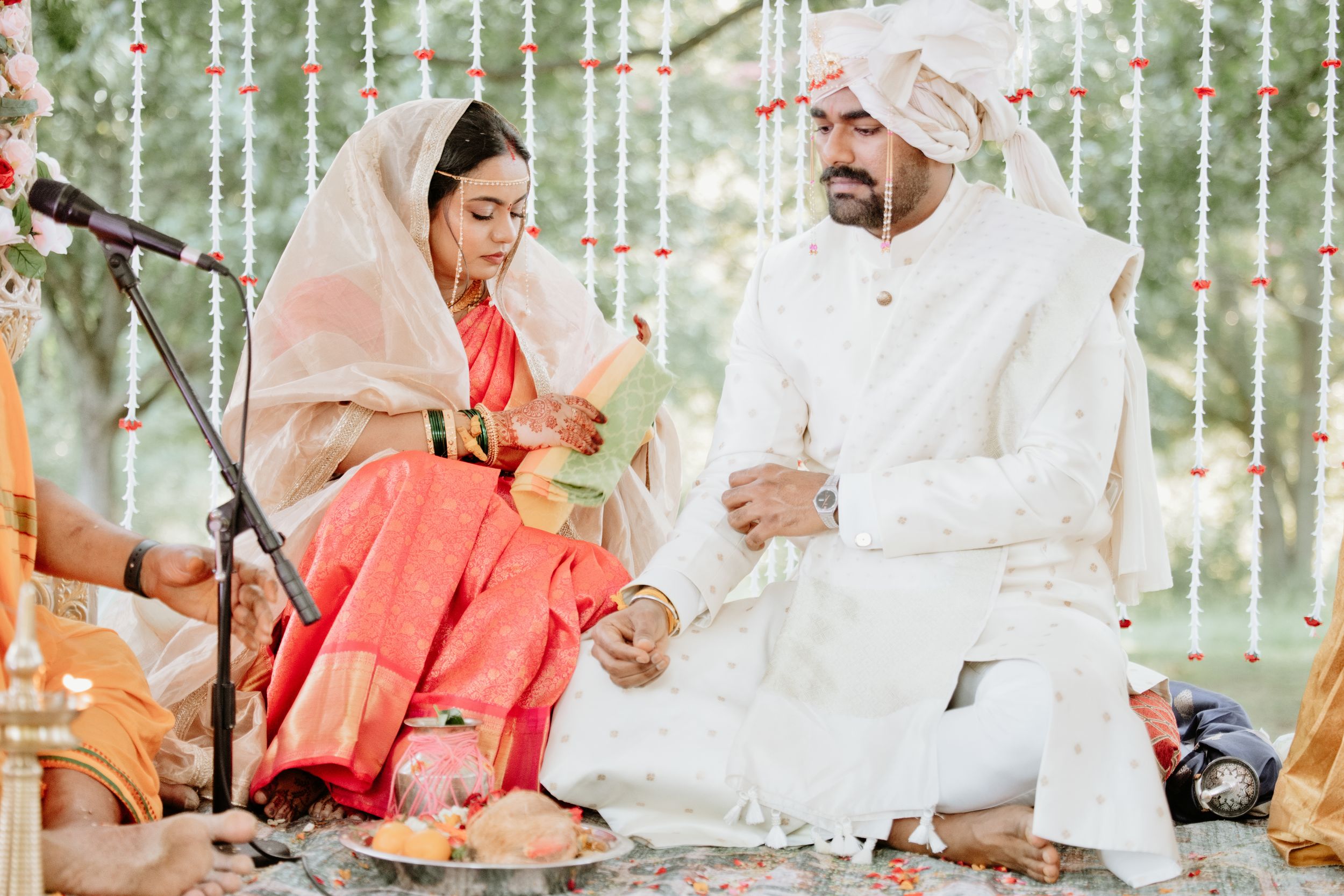 Couple in traditional Indian wedding attire sitting together during a colorful ceremony under floral decorations.