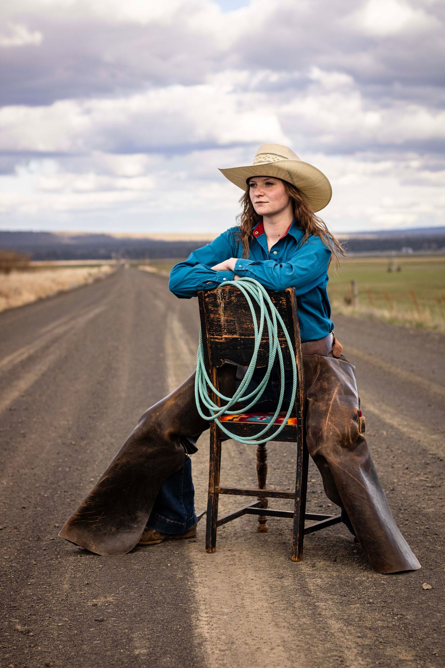 young woman sits on backwards chair with rope, chaps, and cowboy hat. behind her is a long dirt road, hills, and a cloudy sky