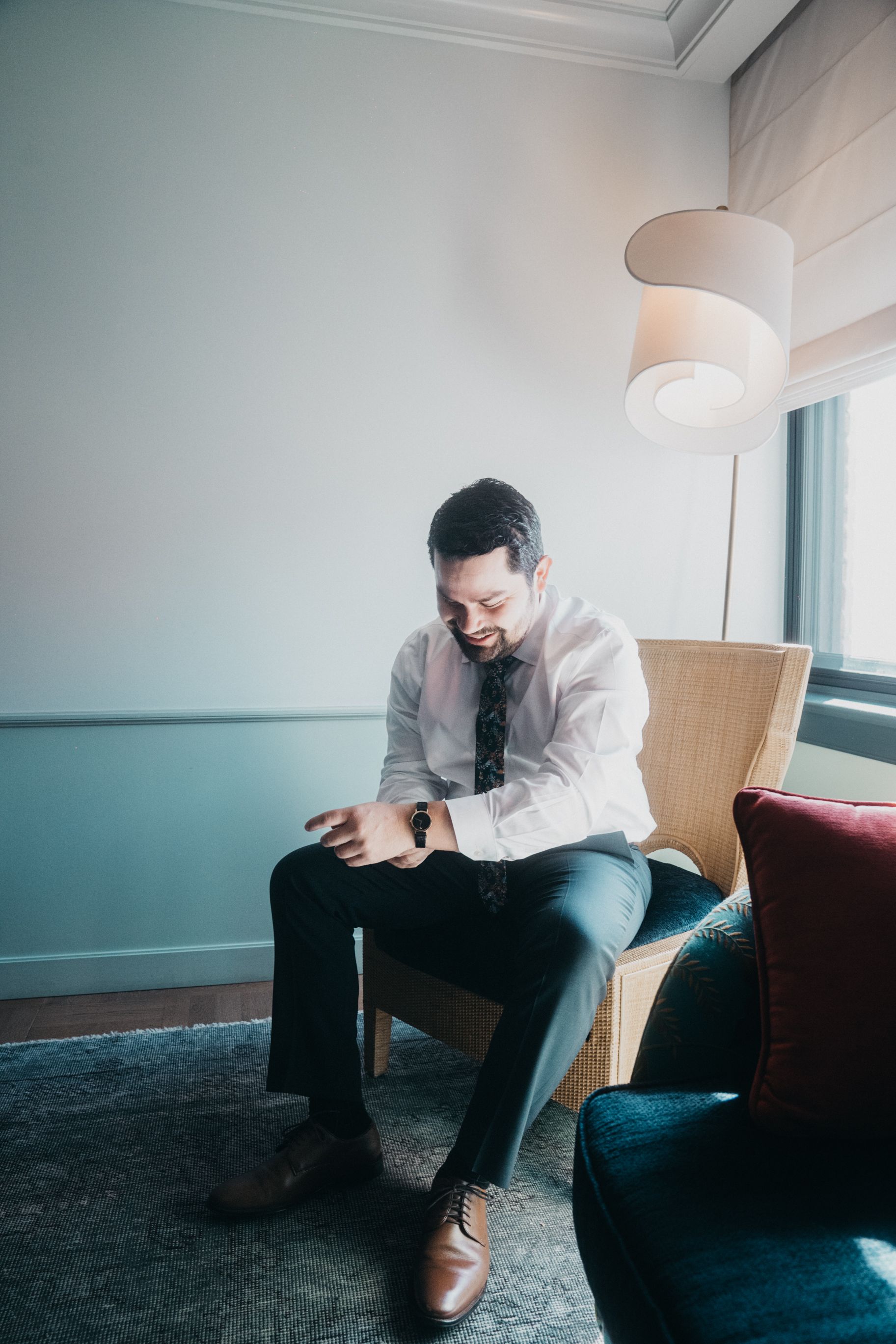 Groom in formal attire checks time while sitting in modern hotel room.