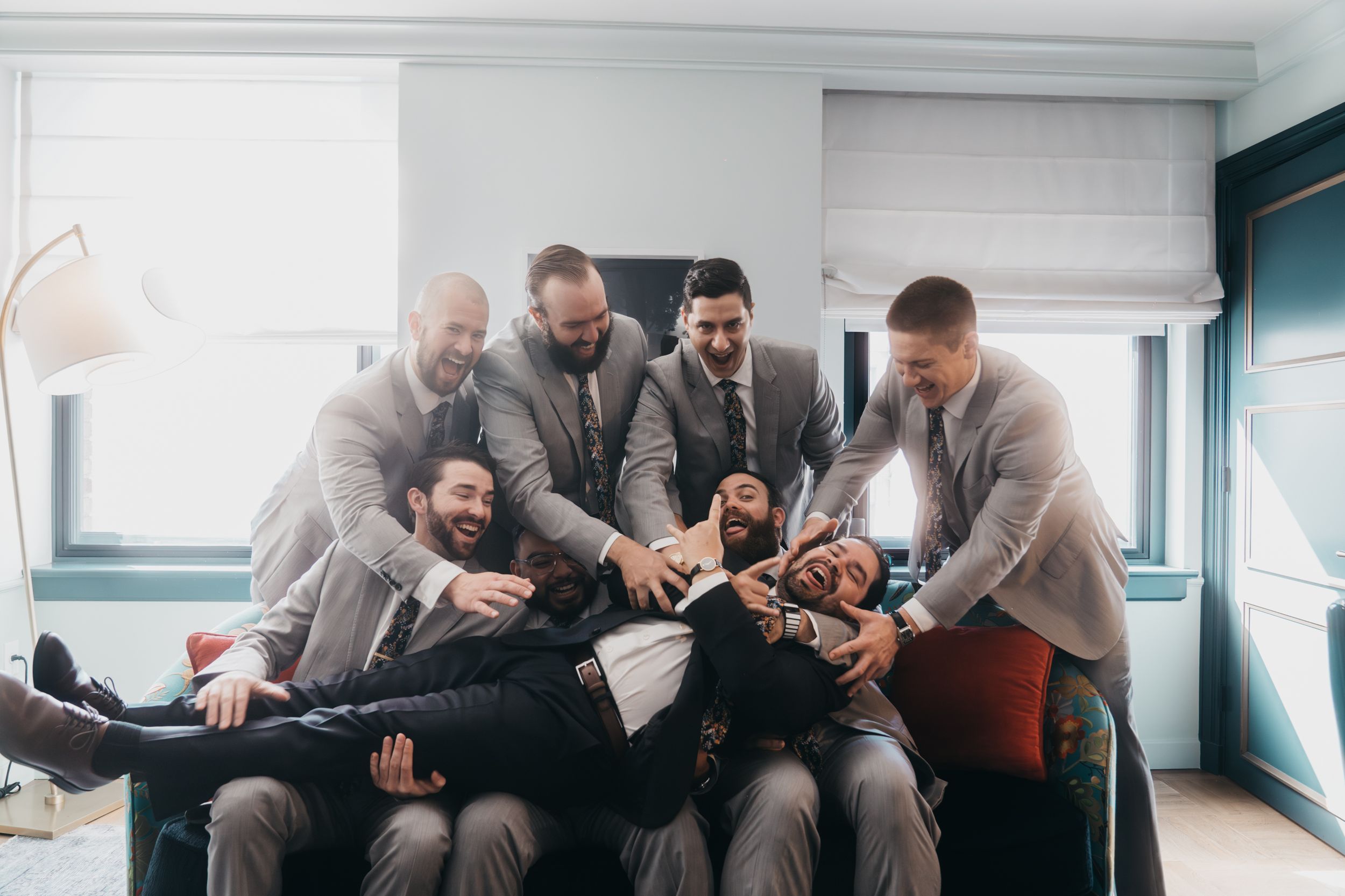 Groomsmen in grey suits sharing drinks and laughs in bright room.