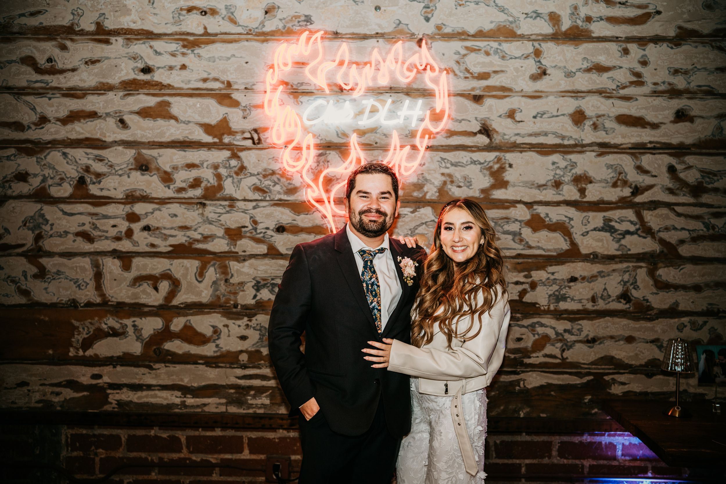 Couple poses against rustic brick wall with glowing neon heart sign.