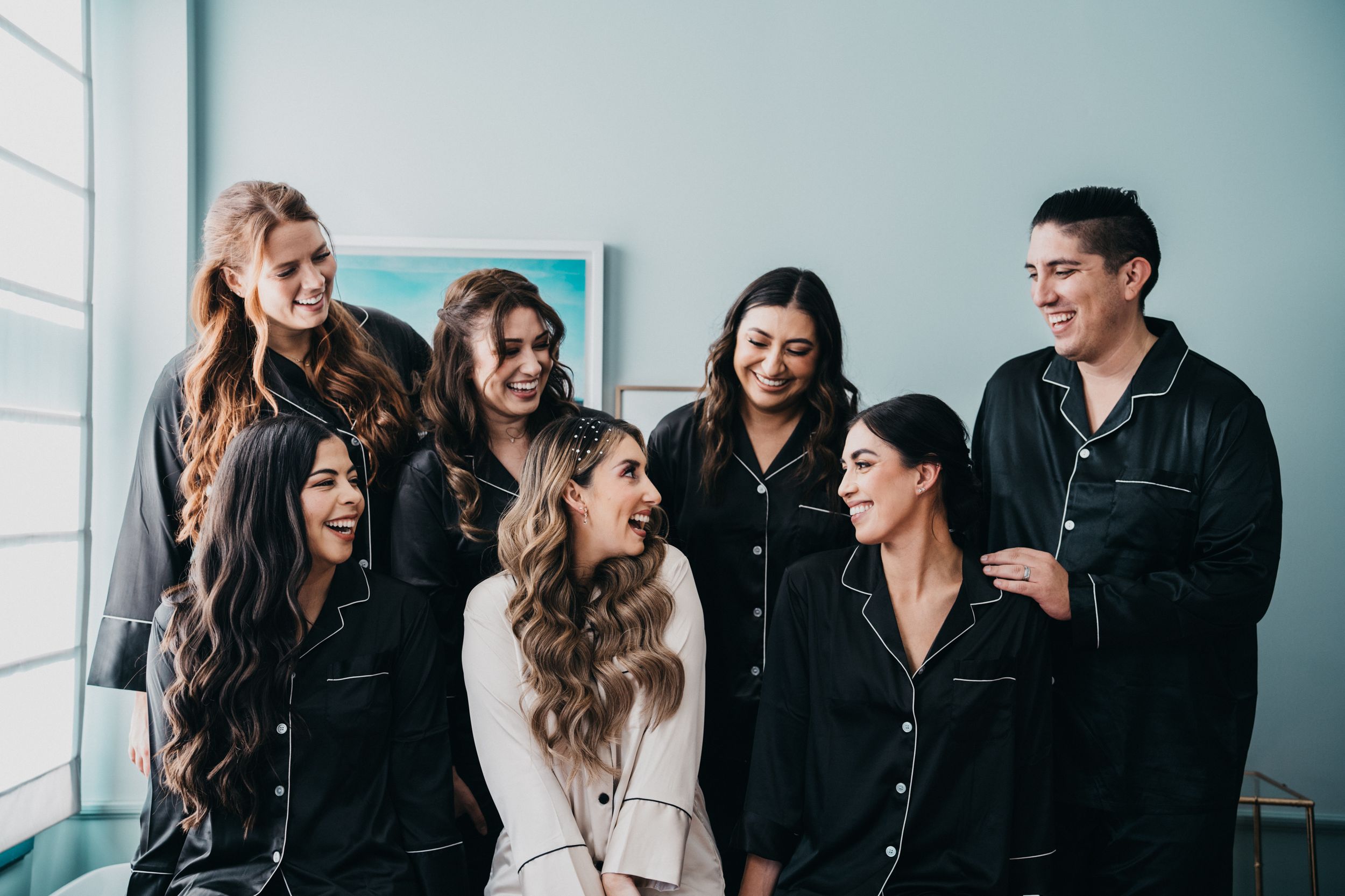 Group in matching black robes sharing laughs by window during pre-wedding preparations.