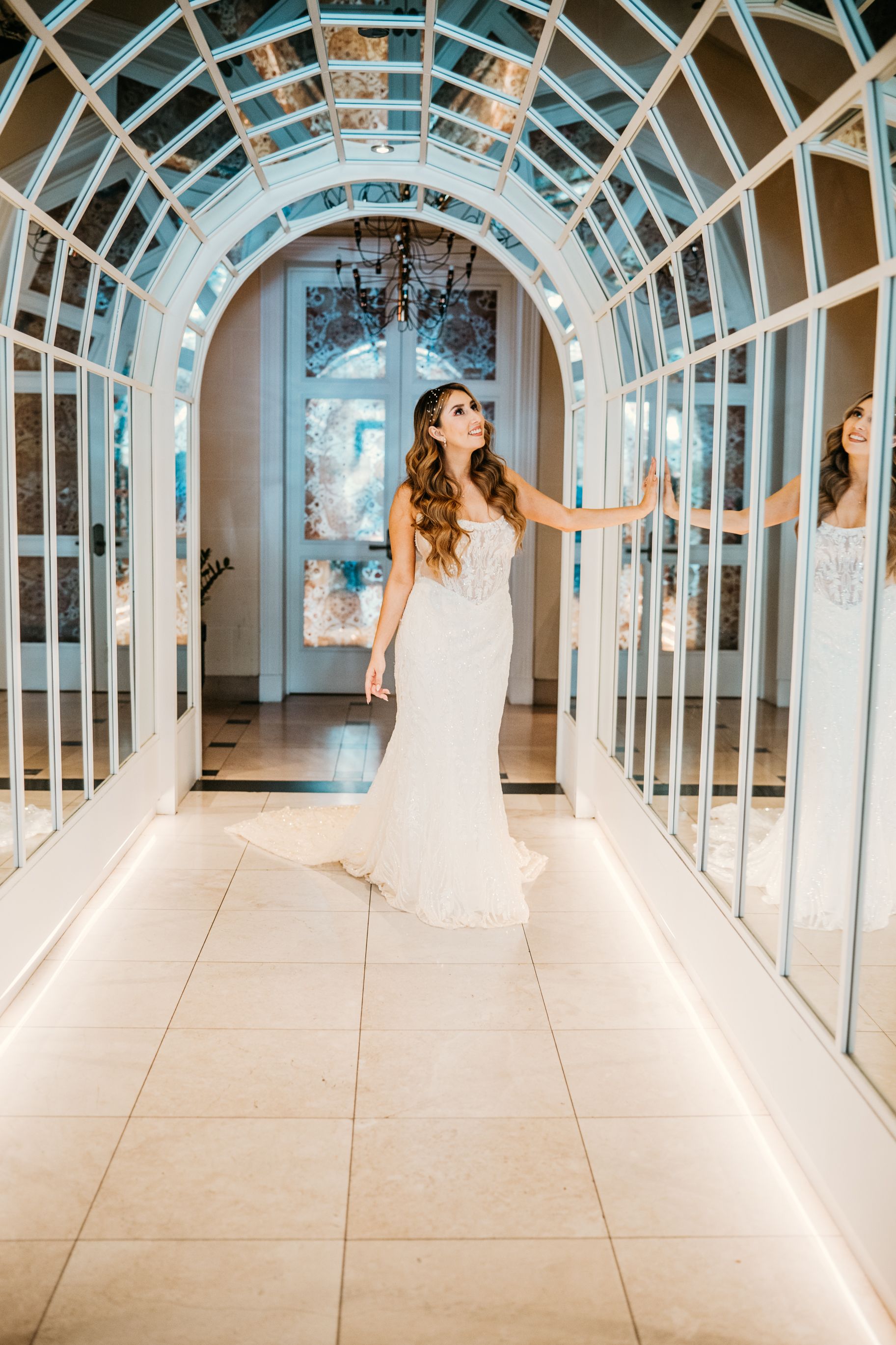 Bride in fitted wedding gown poses in mirrored hallway with arched ceiling.