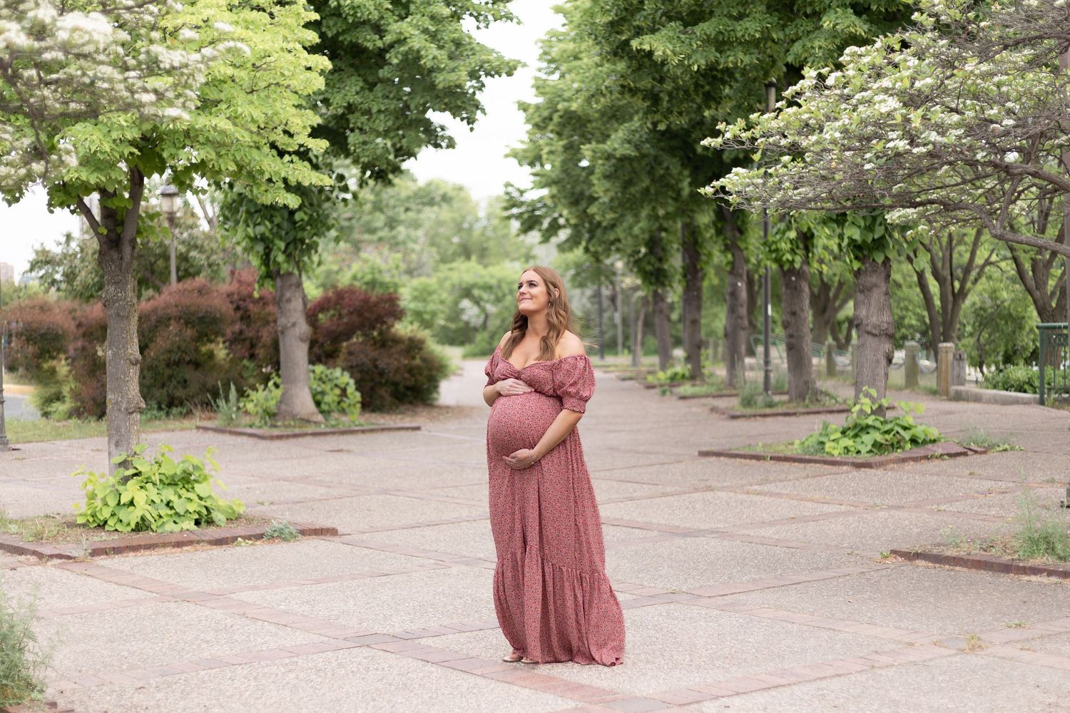 An expectant mother in a flowing mauve dress poses in a tree-lined garden pathway.