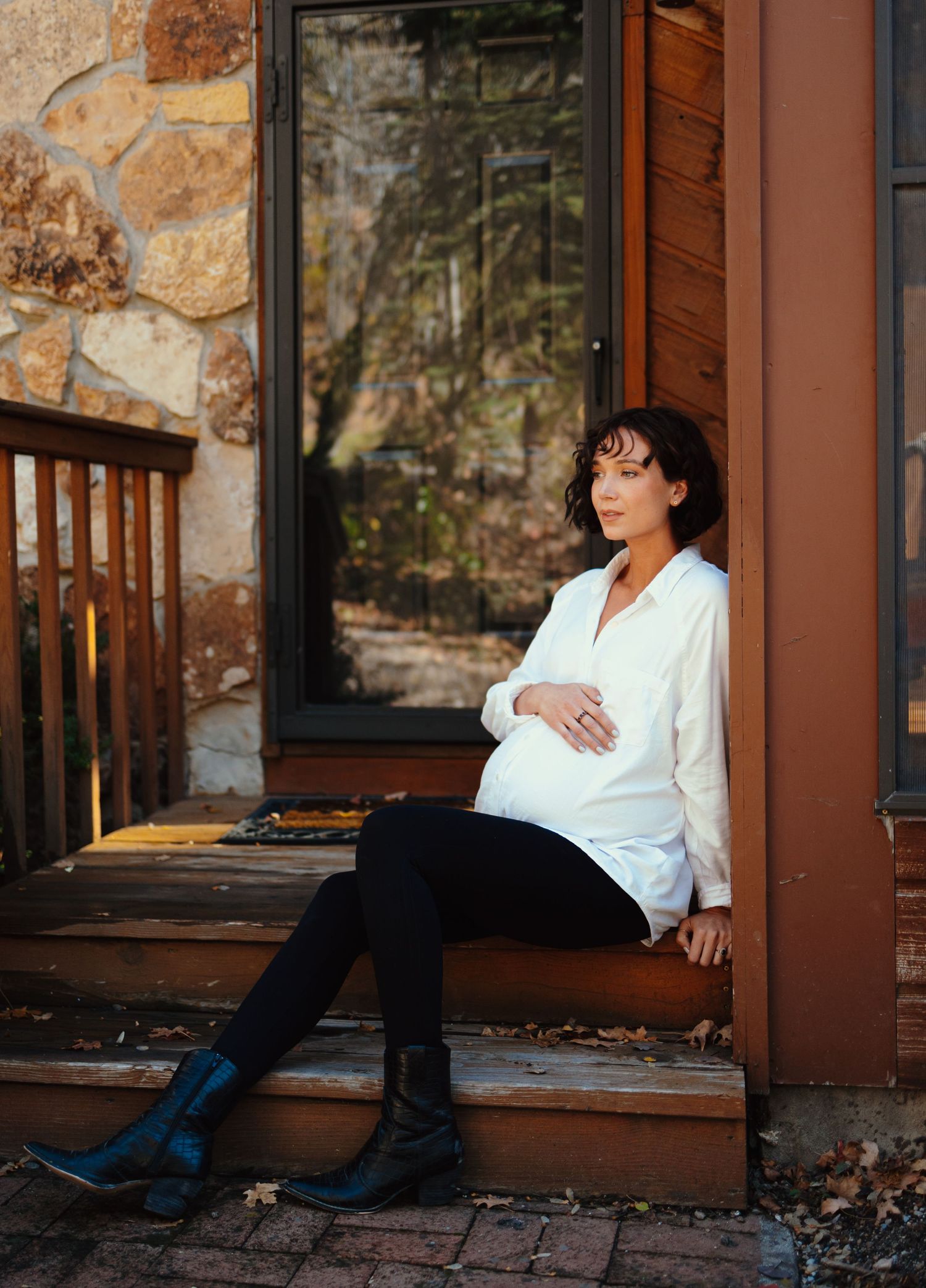 Expectant mother in white shirt and dark pants sits thoughtfully on rustic wooden steps.
