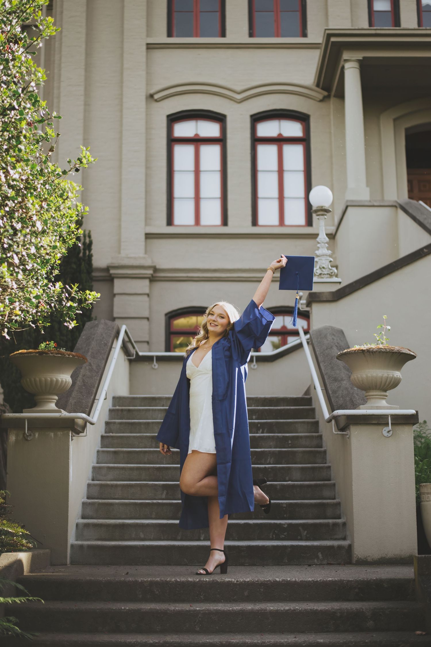 Graduate in blue cap and gown celebrates on stone steps of historic building while holding diploma aloft.