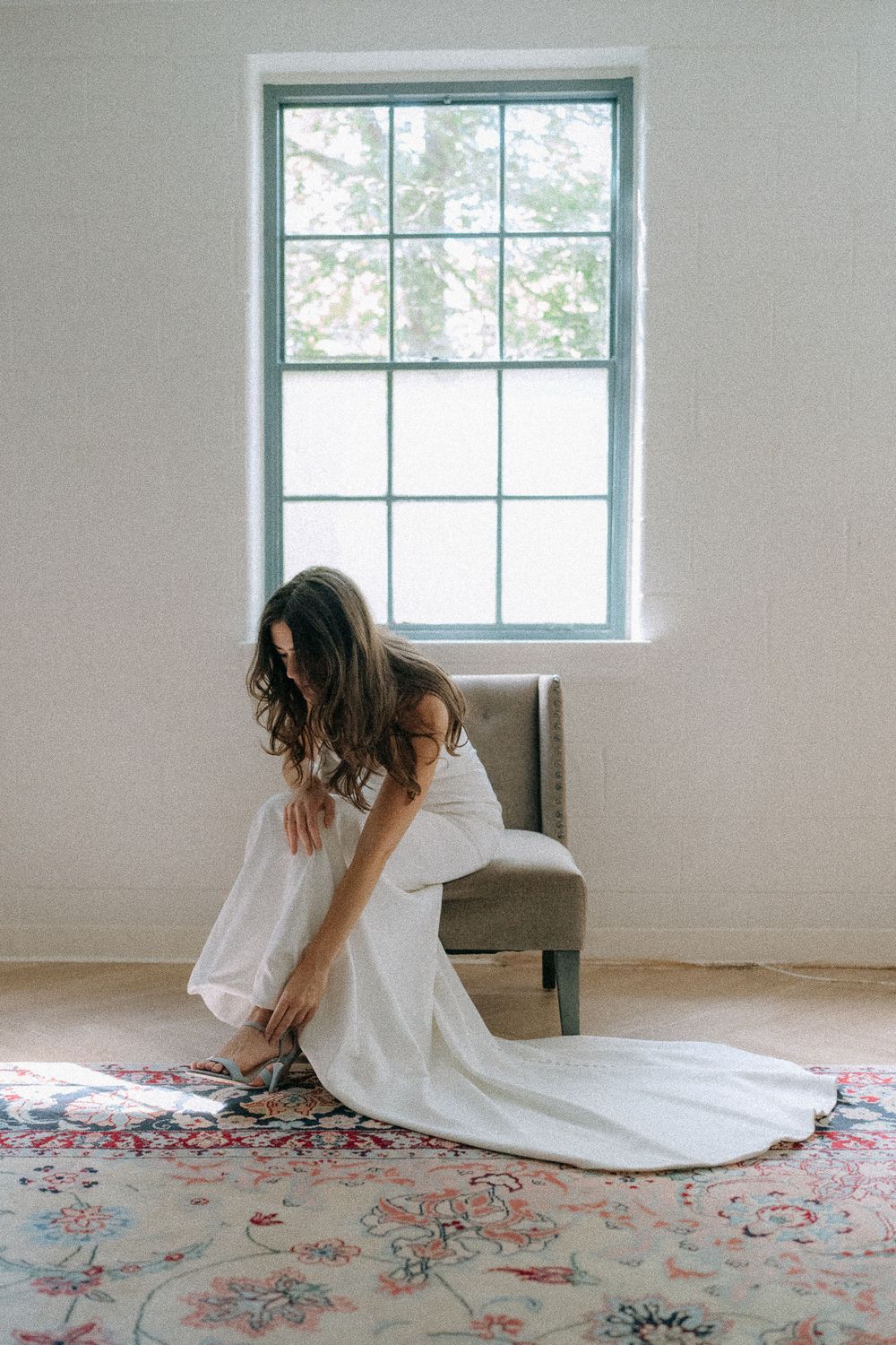 Elegant bride in a white gown adjusting her shoes while seated near a window on a patterned rug.