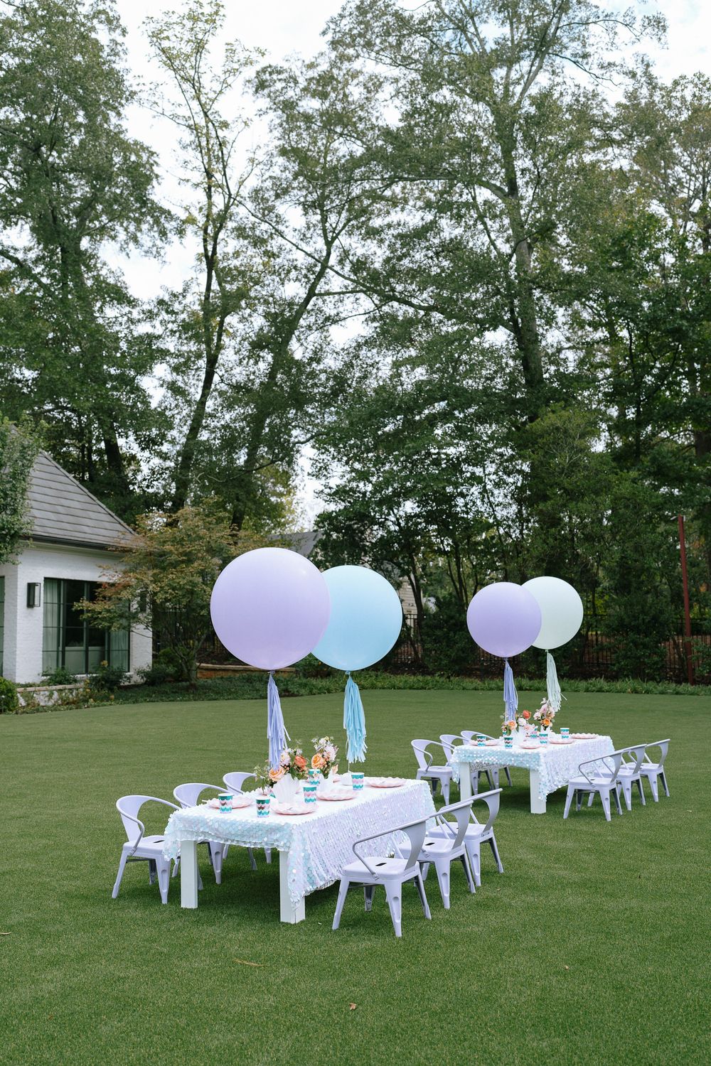 Outdoor party setup with large pastel purple and blue balloons over white tables on green lawn.