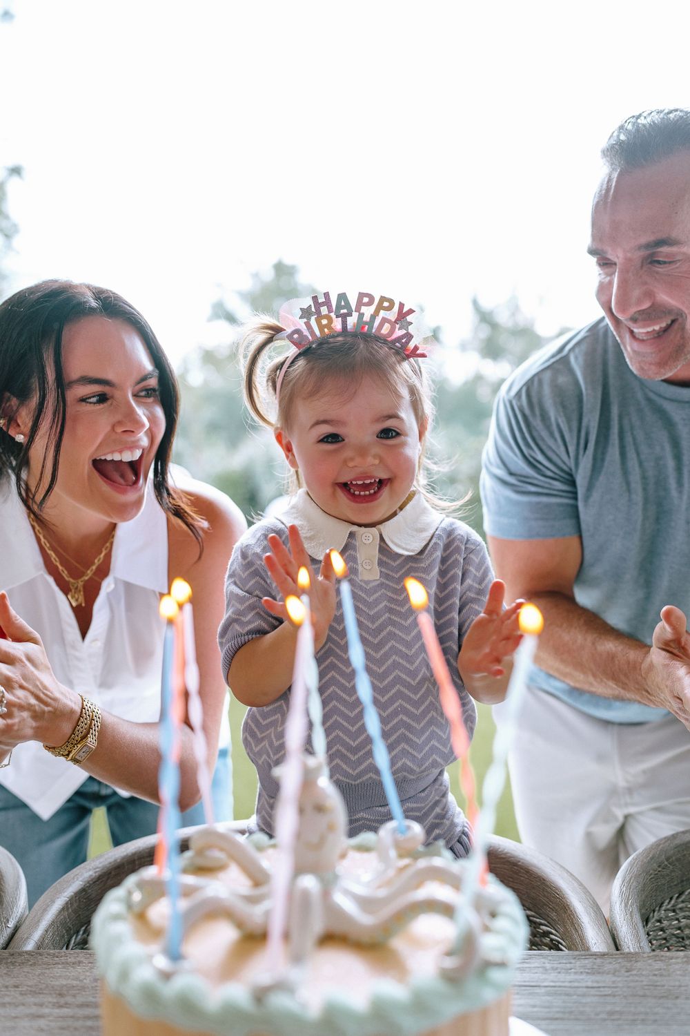 Happy toddler in birthday crown celebrating first birthday with parents and lit candle cake.