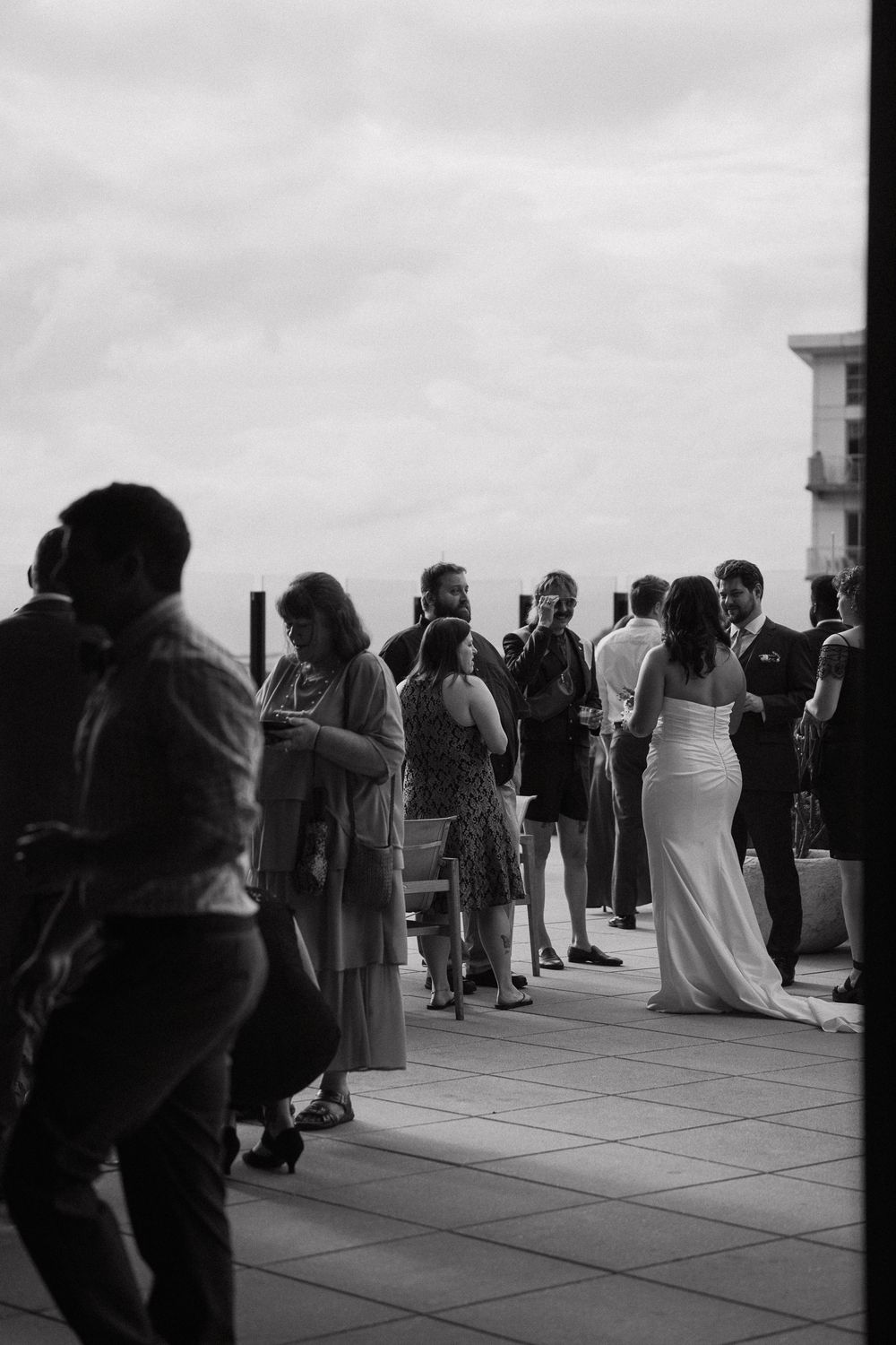 Black and white photo of wedding guests mingling on rooftop terrace with bride in white gown.