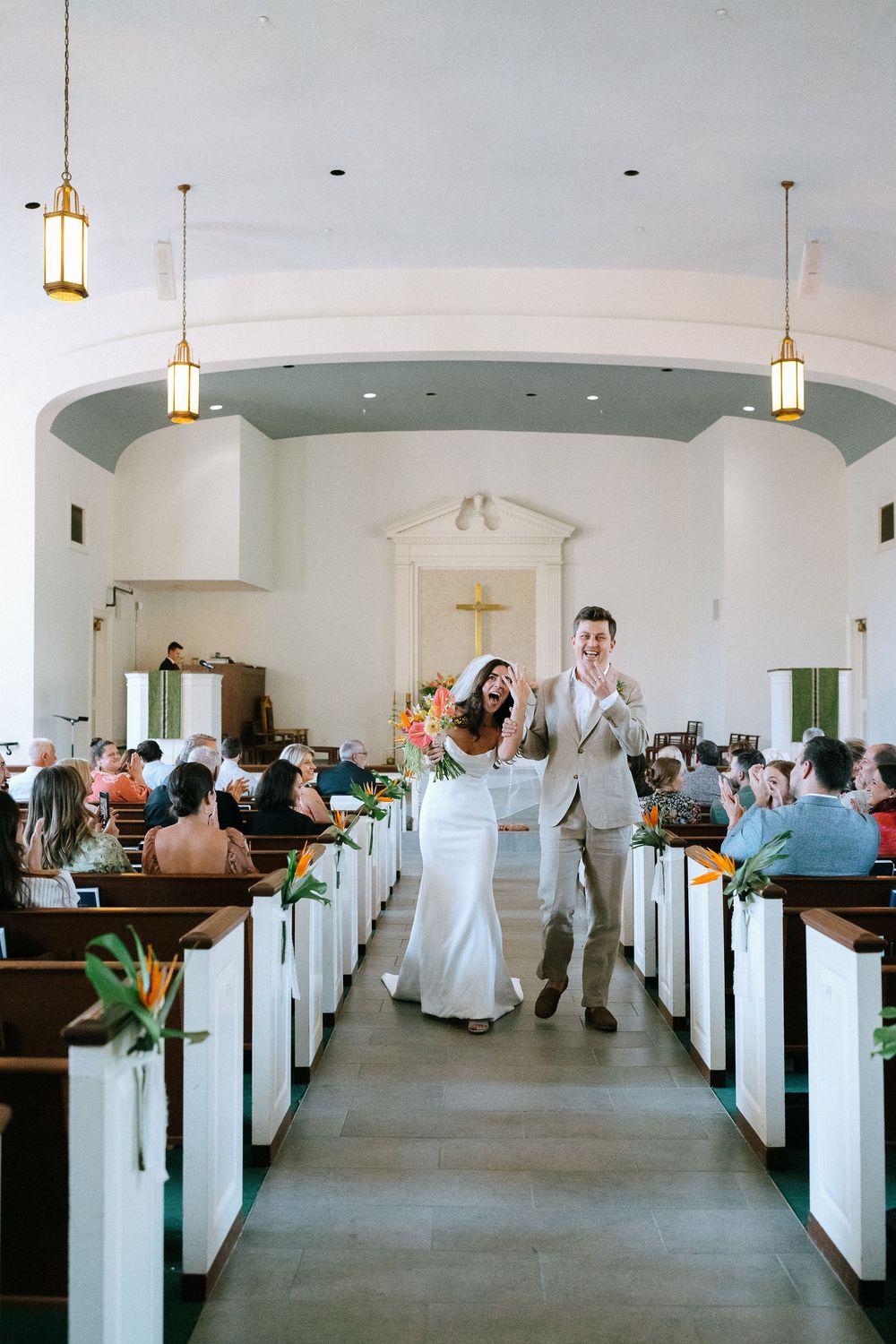 Joyful newlyweds walking down church aisle after wedding ceremony with guests seated in wooden pews.