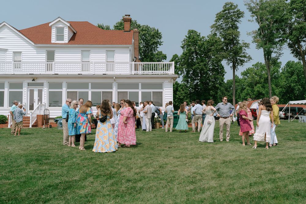 Guests mingling on a green lawn outside a large white colonial house during a sunny outdoor event.