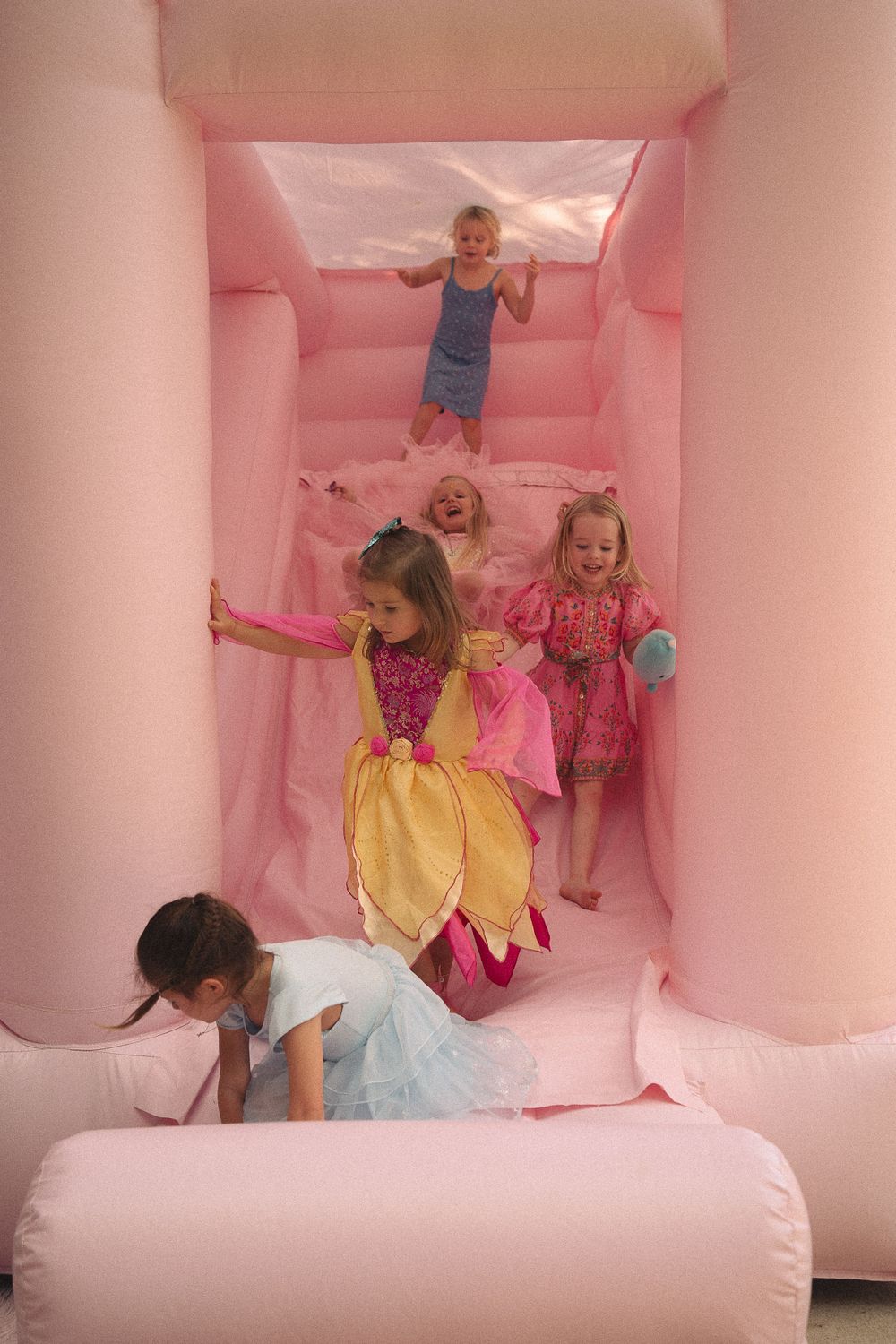 Children in princess costumes laugh and play on a pink bouncy castle slide.