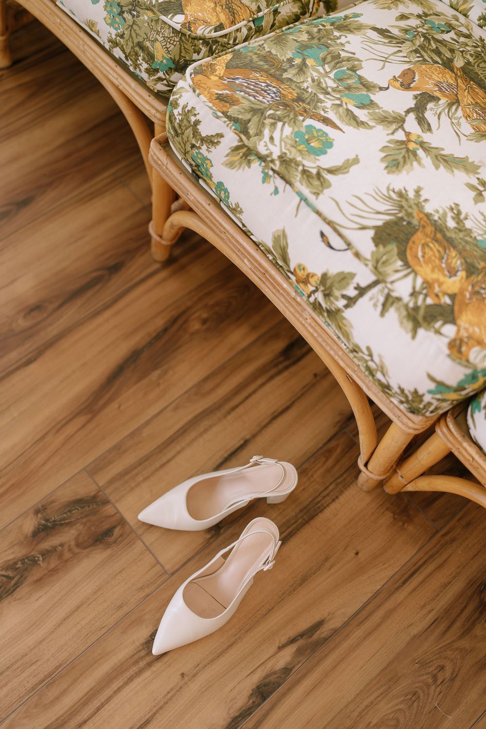 White satin pointed-toe slingback heels resting on hardwood floor beside a rattan chair with floral cushion.