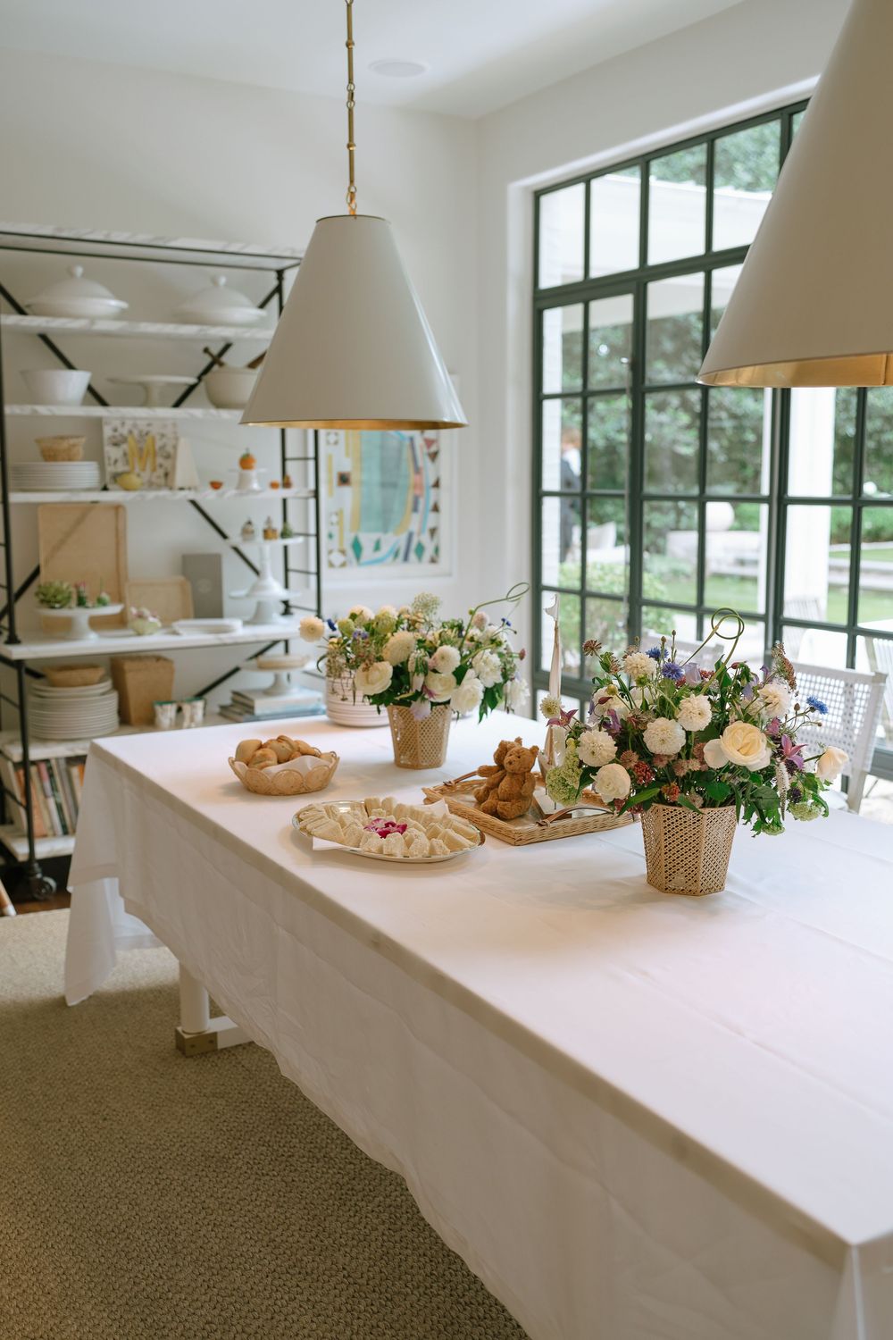 Bright reception table with white linen, floral centerpieces, and pendant lights near large black-framed windows.