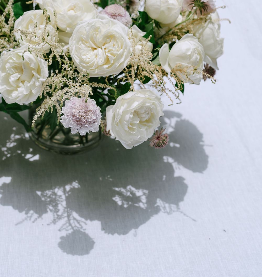 Close-up of white garden roses and soft pink flowers casting delicate shadows on a white linen tablecloth.