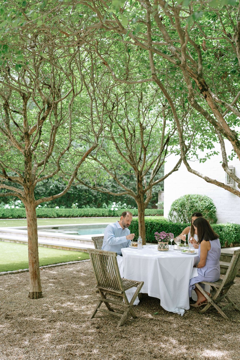 Couple dining al fresco under a canopy of trees beside a pool at an elegant garden estate.