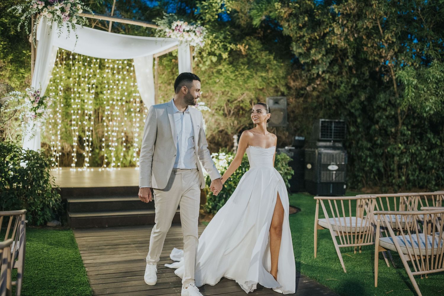 Wedding couple walking near white draped ceremony arch with hanging greenery.