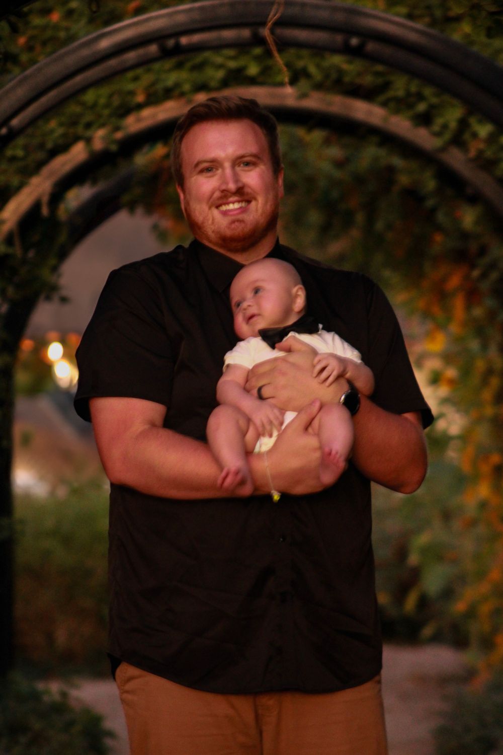 A person in black shirt holds a baby wearing formal attire under an outdoor garden arch at dusk.
