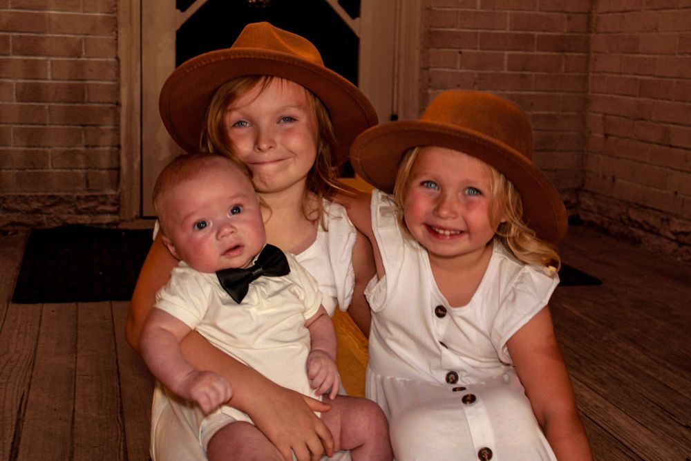 Three children in matching western-style outfits pose together for a nostalgic portrait on wooden floor.