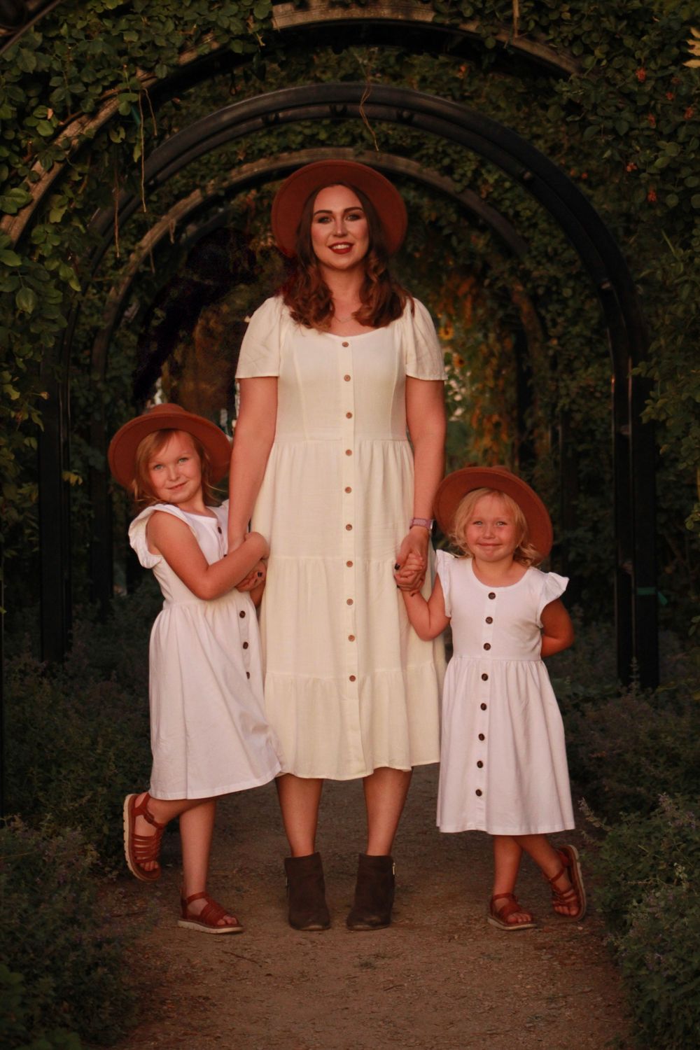 Three people in coordinated white dresses and brown hats pose under a garden archway.