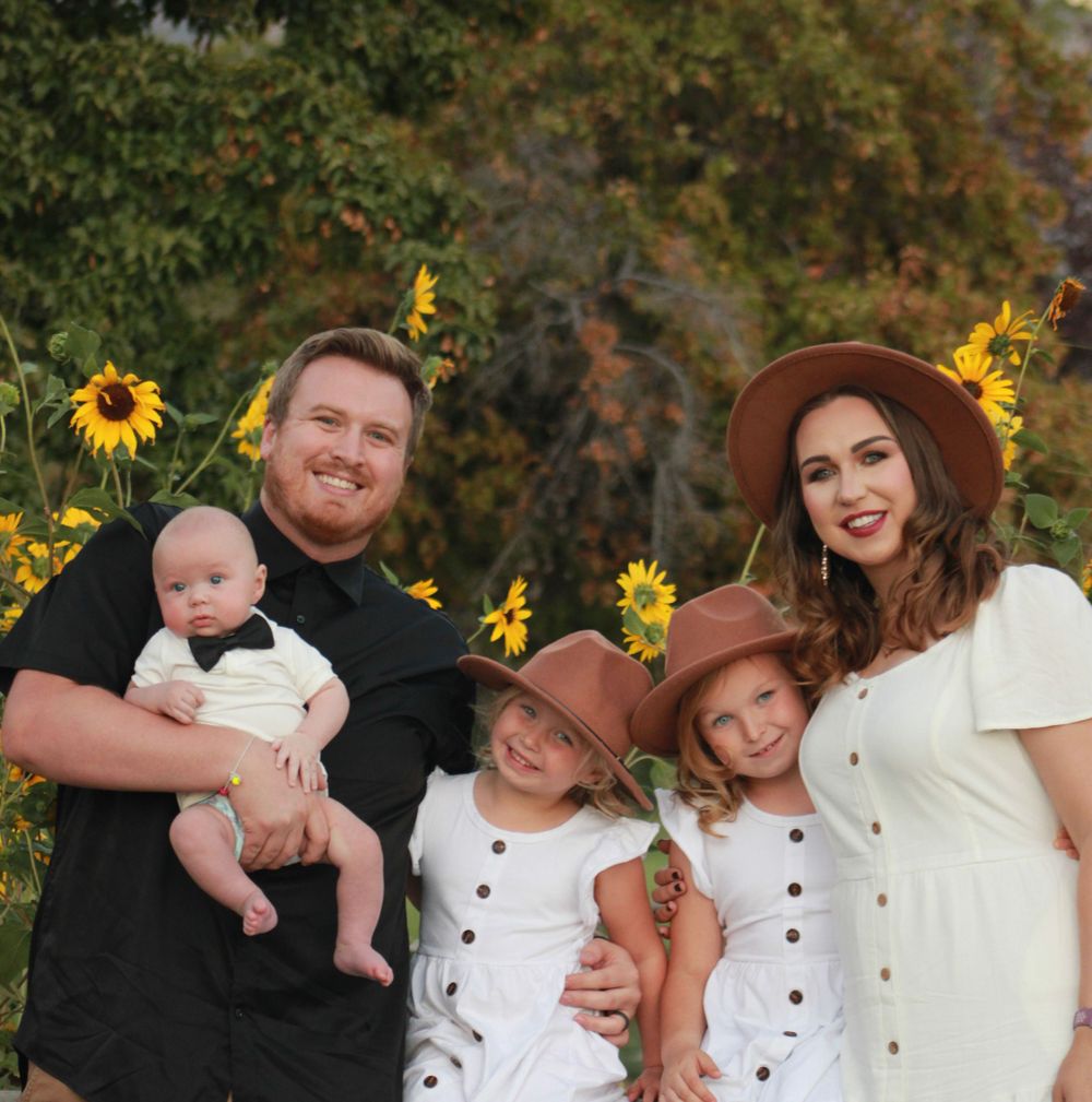 Family poses in front of sunflower field with baby and two children in matching white dresses.