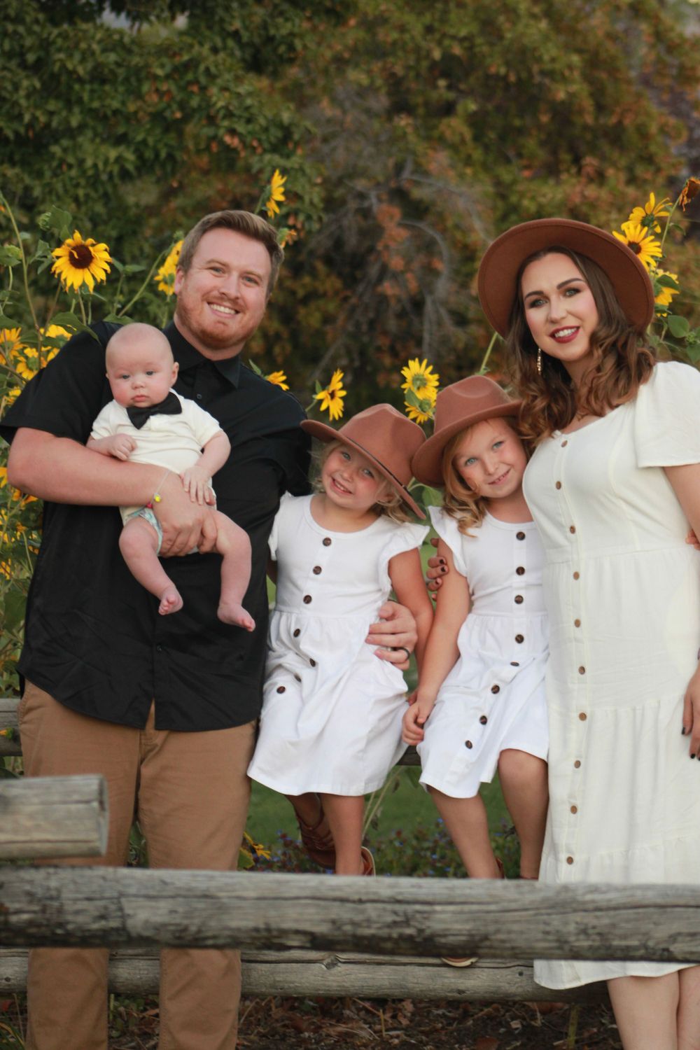 A family poses together in front of sunflowers wearing coordinated white and brown outfits on a rustic fence.