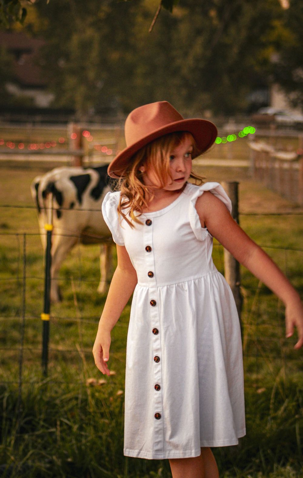 A child in a white button-down dress and brown hat stands near a farm fence.